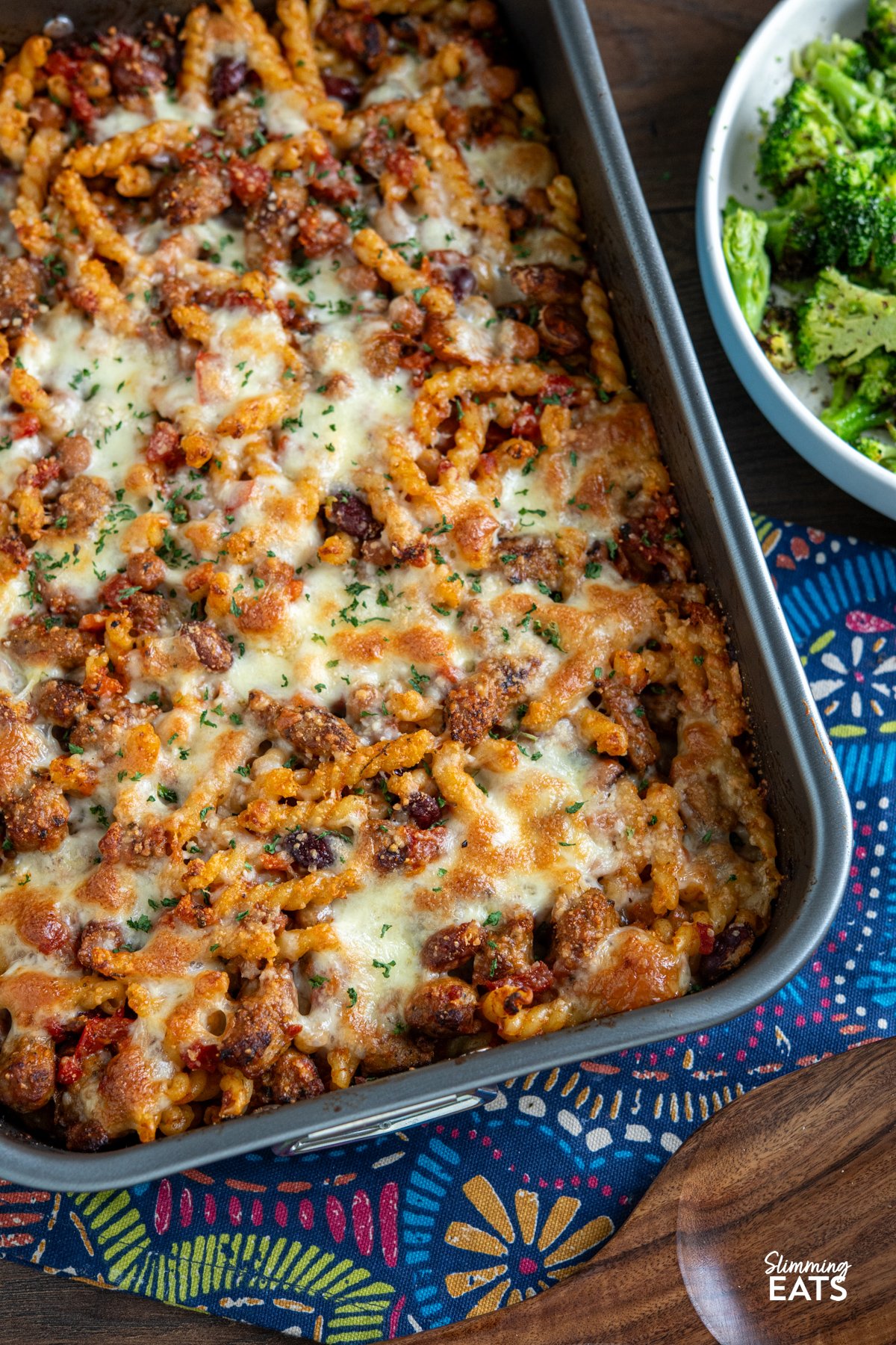 Impress the whole family with this delicious Sausage and Bean Pasta Bake! Packed with all the flavours everyone loves—sausages, beans, and pasta—this dish is both satisfying and simple to make. It's a firm favourite with my kids, who can’t get enough of it, and it’s perfect for both adults and little ones alike. Sausage and Bean Pasta Bake in baking dish on pattern cloth with wooden spatula below and bowl of cooked broccoli to the side