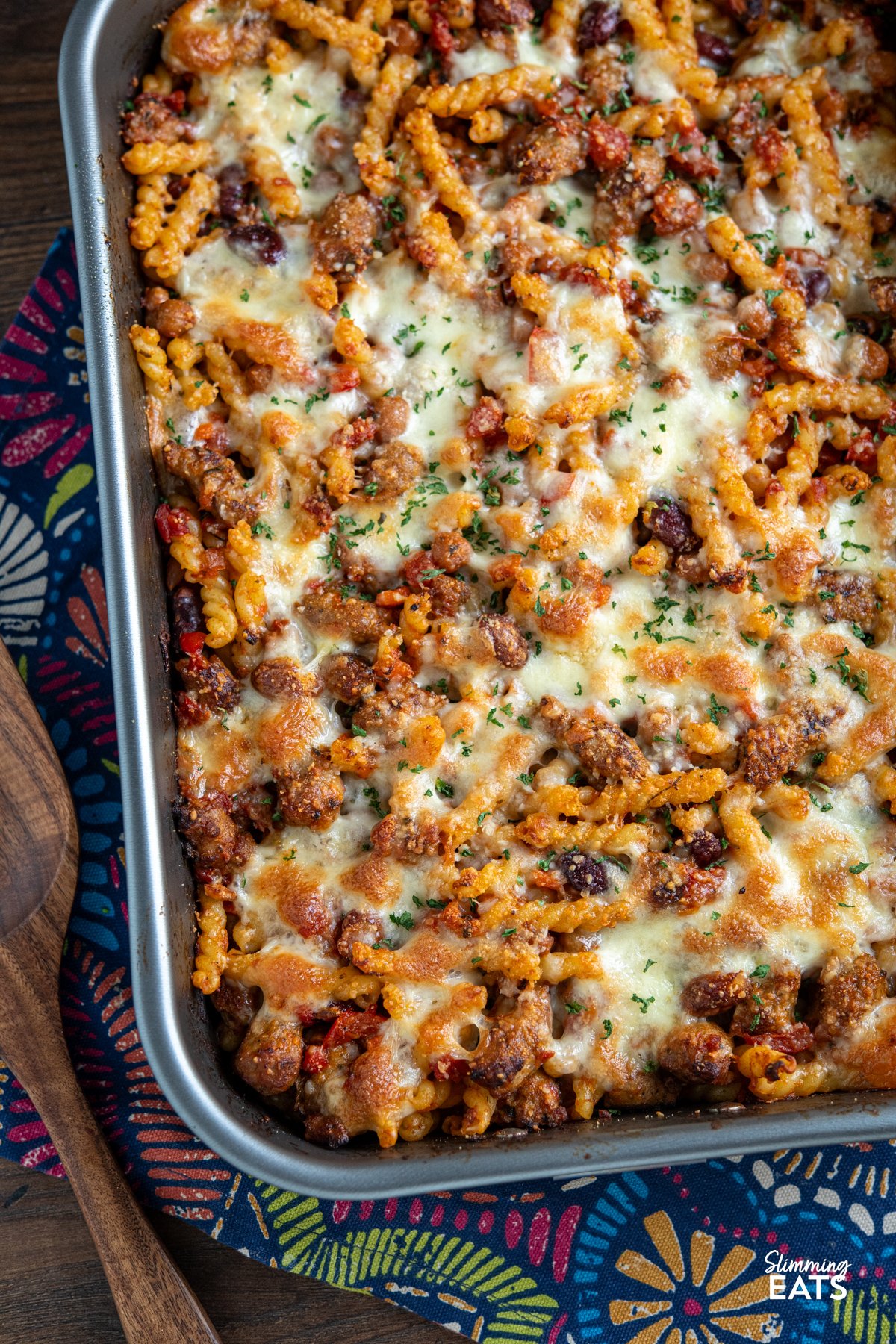 Impress the whole family with this delicious Sausage and Bean Pasta Bake! Packed with all the flavours everyone loves—sausages, beans, and pasta—this dish is both satisfying and simple to make. It's a firm favourite with my kids, who can’t get enough of it, and it’s perfect for both adults and little ones alike. close up of Sausage and Bean Pasta Bake in baking dish on pattern cloth with wooden spatula to side