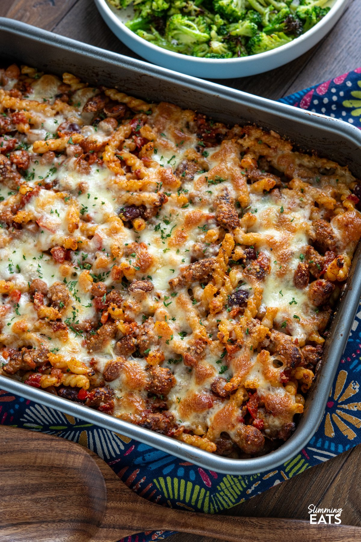 Impress the whole family with this delicious Sausage and Bean Pasta Bake! Packed with all the flavours everyone loves—sausages, beans, and pasta—this dish is both satisfying and simple to make. It's a firm favourite with my kids, who can’t get enough of it, and it’s perfect for both adults and little ones alike. close up of Sausage and Bean Pasta Bake in baking dish on pattern cloth with wooden spatula below and bowl of cooked broccoli above.