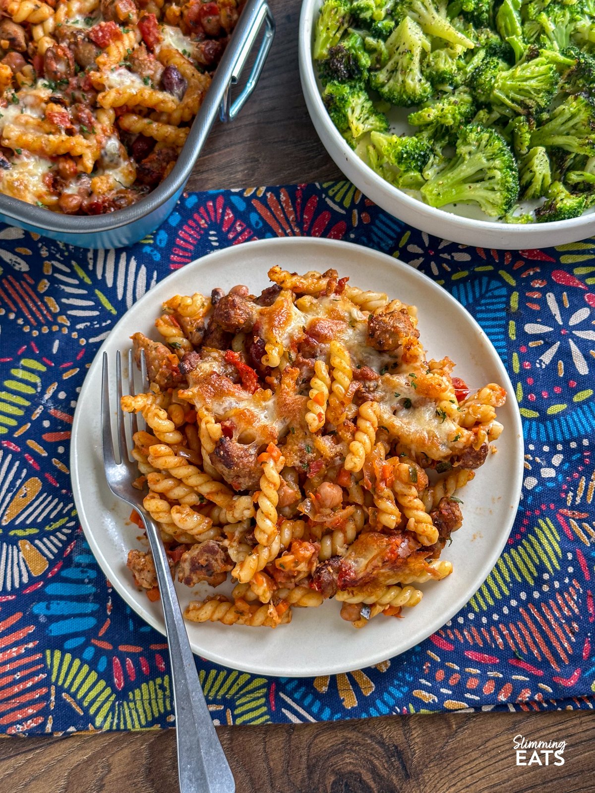 Impress the whole family with this delicious Sausage and Bean Pasta Bake! Packed with all the flavours everyone loves—sausages, beans, and pasta—this dish is both satisfying and simple to make. It's a firm favourite with my kids, who can’t get enough of it, and it’s perfect for both adults and little ones alike. a portion of Sausage and Bean Pasta Bake served up on a white plate with fork to the side, dish of pasta bake and bowl of cooked broccoli in background