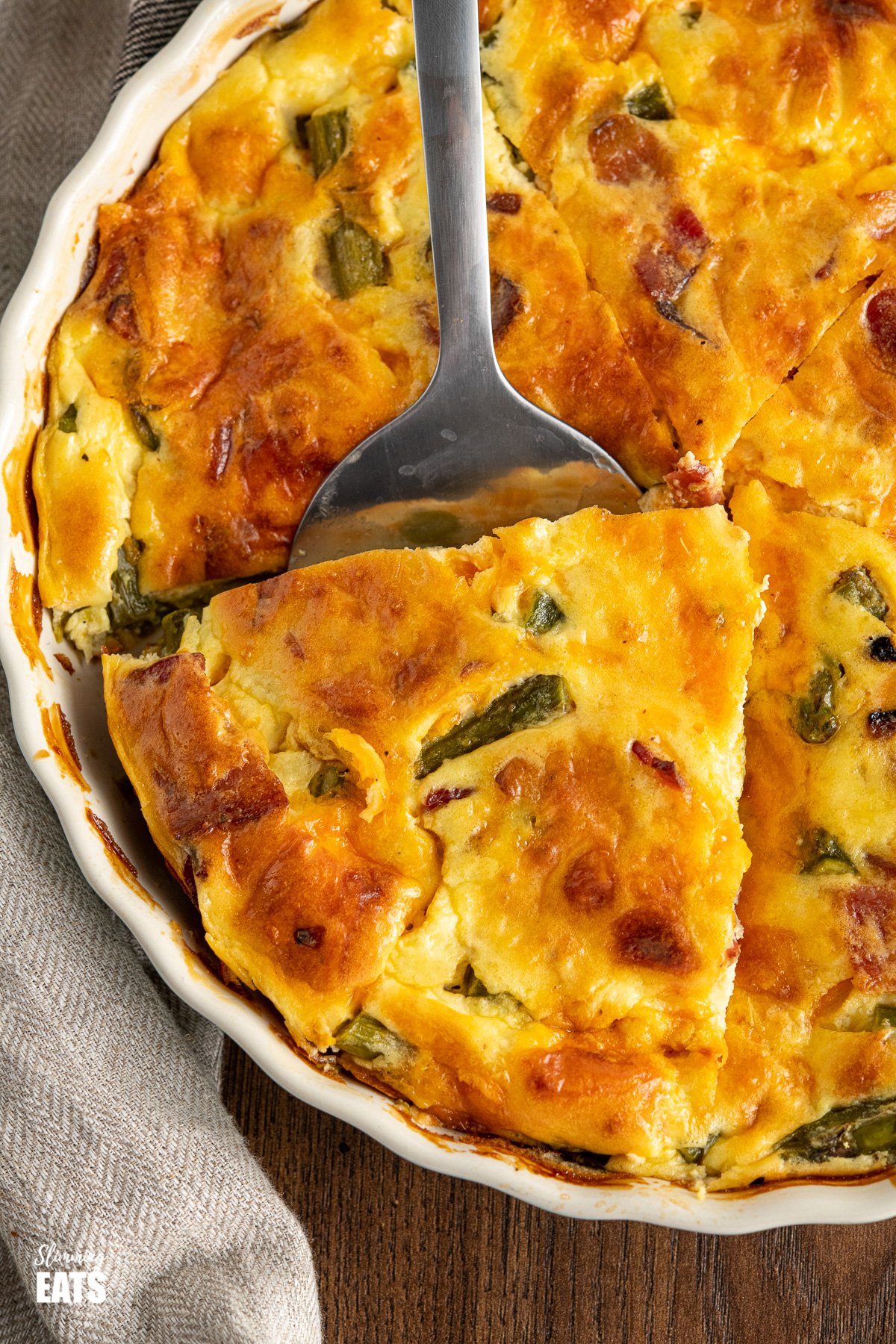 close up of slice of Crustless Asparagus and Bacon Quiche in flan dish with spatula on wooden board