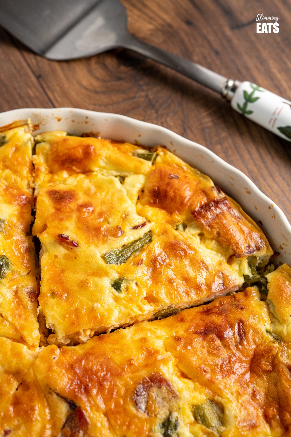 close up of slice of Crustless Asparagus and Bacon Quiche in flan dish with spatula on wooden board