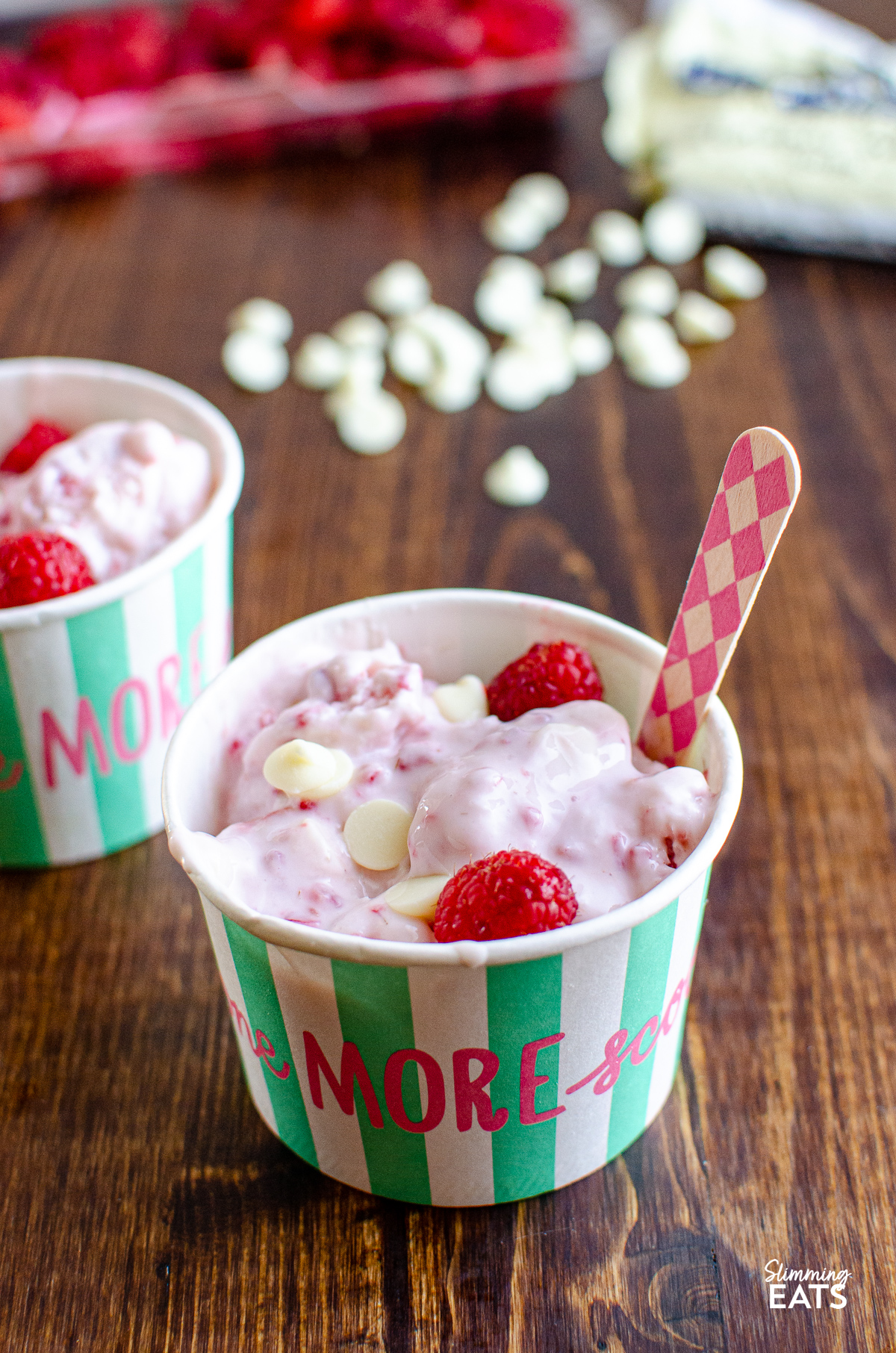 Raspberry and White Chocolate Frozen Yoghurt in a icecream tub with pink diamond patterned wooden spoon, and scattered white chocolate chips in background