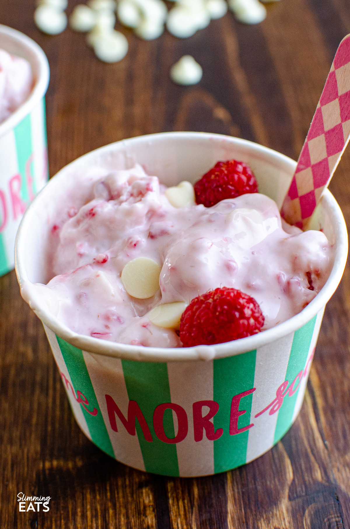 close up of Raspberry and White Chocolate Frozen Yoghurt in a icecream tub with pink diamond patterned wooden spoon, and scattered white chocolate chips in background