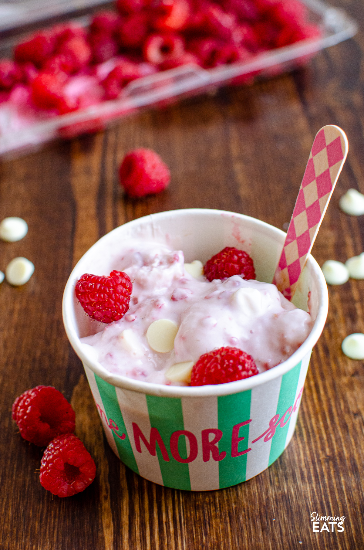 Raspberry and White Chocolate Frozen Yoghurt in a icecream tub with pink diamond patterned wooden spoon, and fresh raspberries  in background.