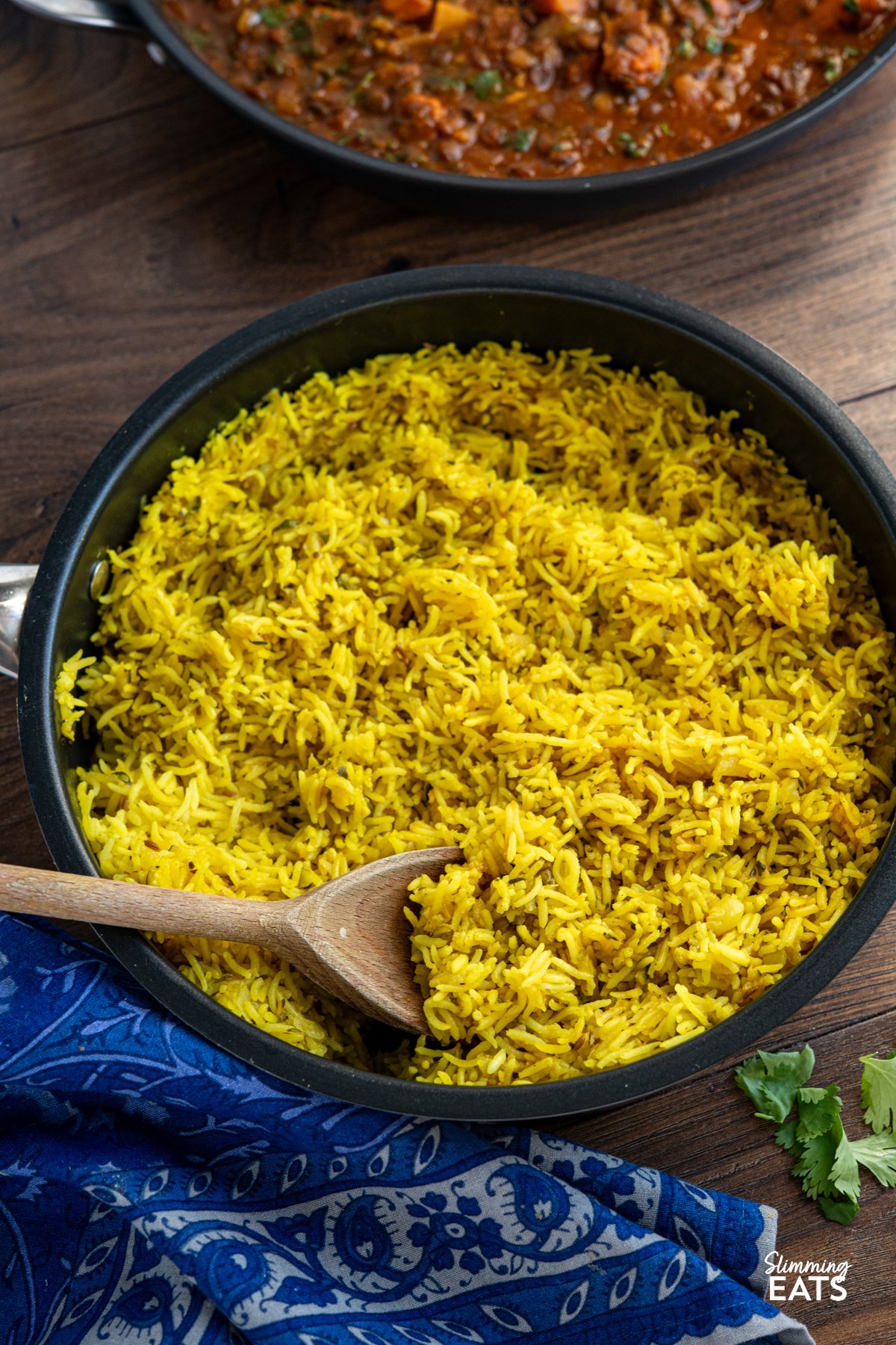 Zucchini Pilau Rice is a fragrant blend of basmati rice, tender zucchini, and warming spices like cumin and turmeric. Light yet flavourful, it’s perfect for a quick dinner or as a vibrant side dish. Zucchini Pilau Rice in a non stick frying pan with wooden spoon, blue patterned napkin underneath and some scattered coriander.
