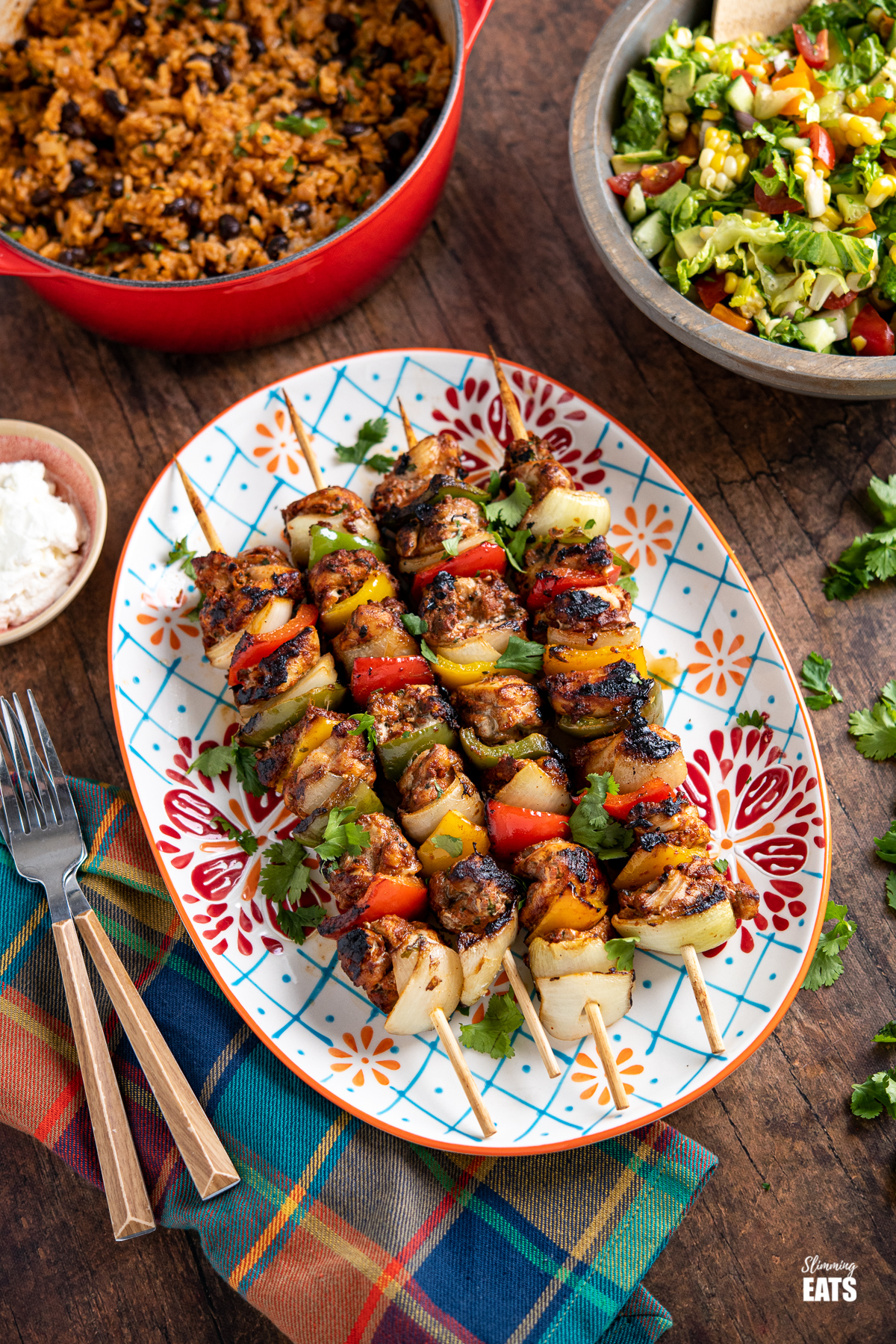 chicken skewers on a oval patterned plate with a pan or rice and bowl of salad in background