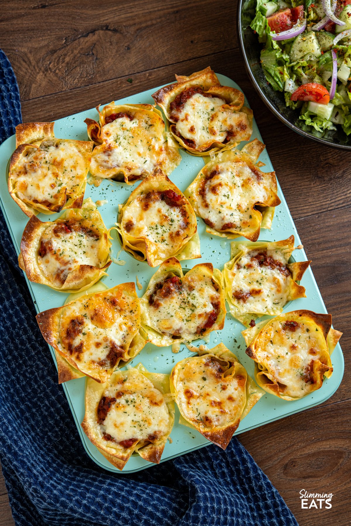 These Mini Lasagne Cups take all the best bits of a classic lasagne and reimagine them into perfectly portioned bites. Instead of layers, cooked lasagne sheets are shaped into little cups and stuffed with a rich, speedy bolognese. Topped with a light béchamel-style sauce and a scattering of cheese, they bake into golden-topped bundles of comfort the whole family will love. Mini Lasagne Cups in a blue muffin tray with a bowl of salad to the right.