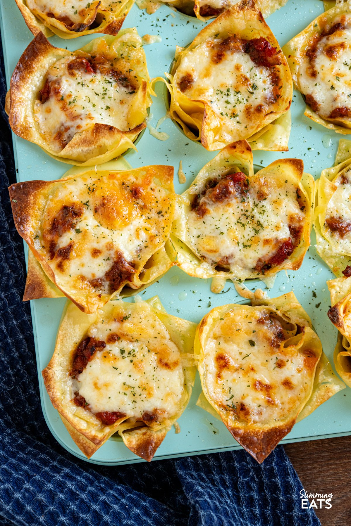 These Mini Lasagne Cups take all the best bits of a classic lasagne and reimagine them into perfectly portioned bites. Instead of layers, cooked lasagne sheets are shaped into little cups and stuffed with a rich, speedy bolognese. Topped with a light béchamel-style sauce and a scattering of cheese, they bake into golden-topped bundles of comfort the whole family will love. close up of Mini Lasagne Cups in a blue muffin tray