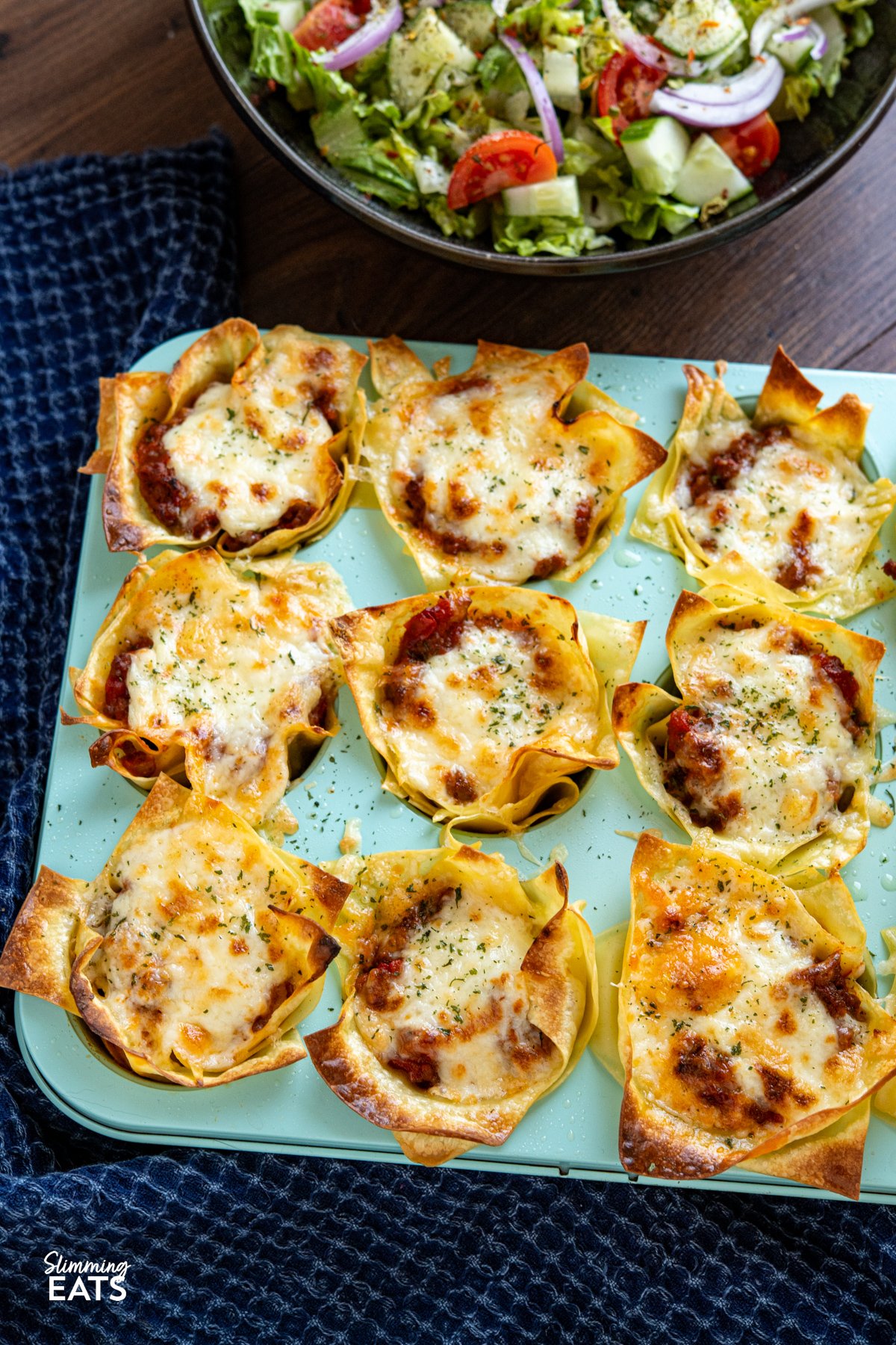 These Mini Lasagne Cups take all the best bits of a classic lasagne and reimagine them into perfectly portioned bites. Instead of layers, cooked lasagne sheets are shaped into little cups and stuffed with a rich, speedy bolognese. Topped with a light béchamel-style sauce and a scattering of cheese, they bake into golden-topped bundles of comfort the whole family will love. Mini Lasagne Cups in a blue muffin tray with a bowl of salad above.