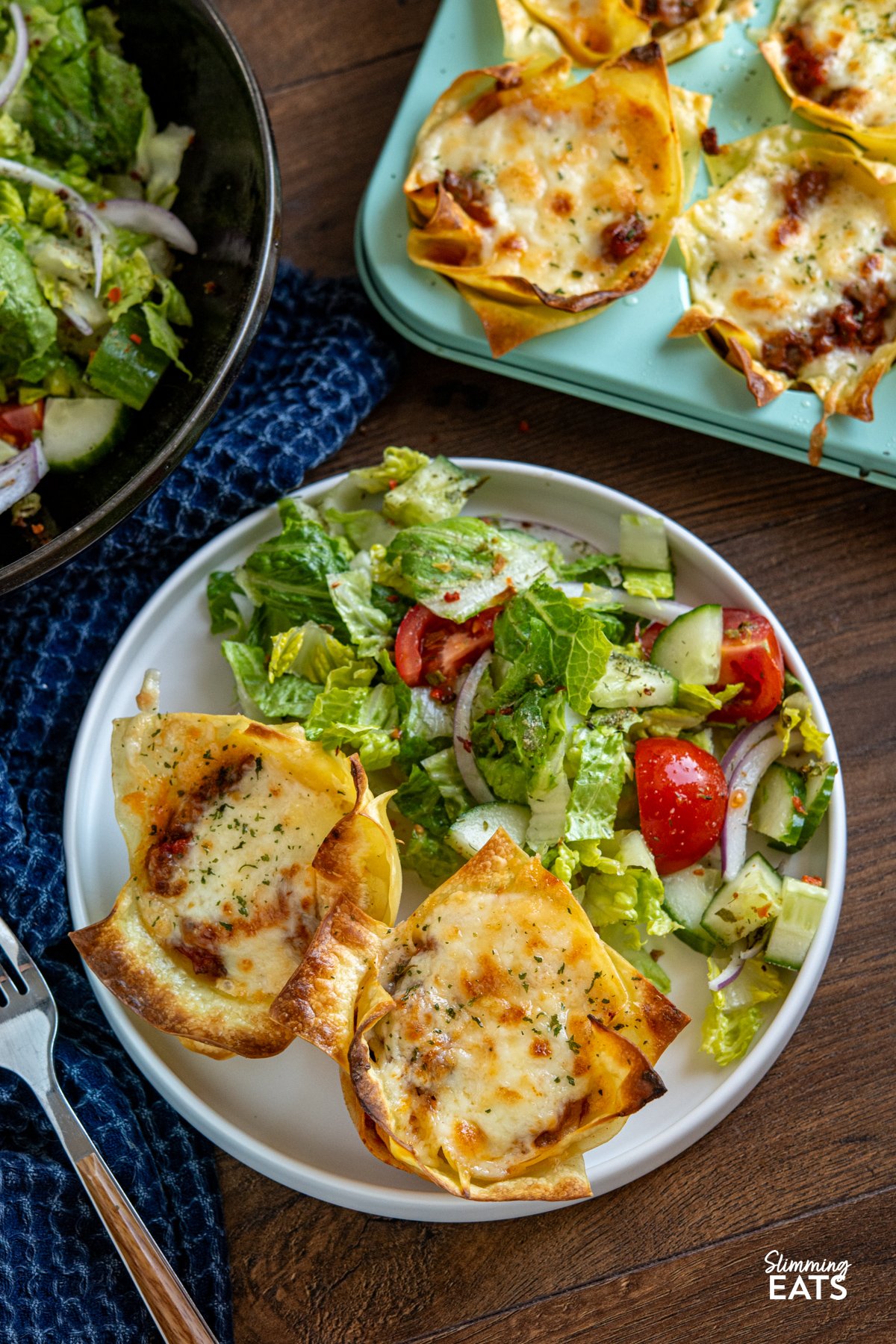These Mini Lasagne Cups take all the best bits of a classic lasagne and reimagine them into perfectly portioned bites. Instead of layers, cooked lasagne sheets are shaped into little cups and stuffed with a rich, speedy bolognese. Topped with a light béchamel-style sauce and a scattering of cheese, they bake into golden-topped bundles of comfort the whole family will love. 2 mini lasagne cups on a white plate with a mixed salad, fork placed to left, muffin tray and bowl of mixed salad visible above,
