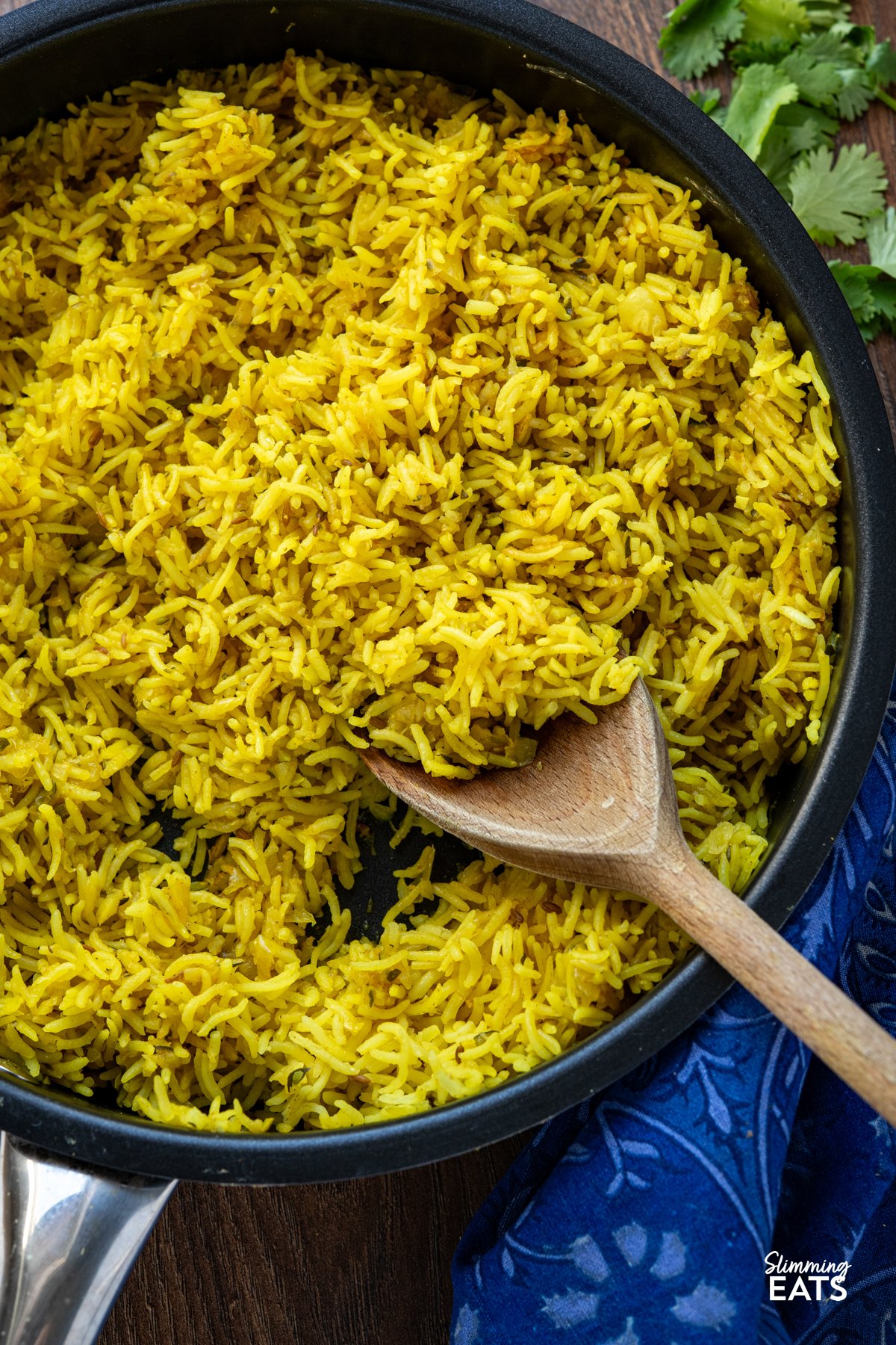Zucchini Pilau Rice is a fragrant blend of basmati rice, tender zucchini, and warming spices like cumin and turmeric. Light yet flavourful, it’s perfect for a quick dinner or as a vibrant side dish. Zucchini Pilau Rice in a non stick frying pan with wooden spoon, blue patterned napkin underneath and some scattered coriander.