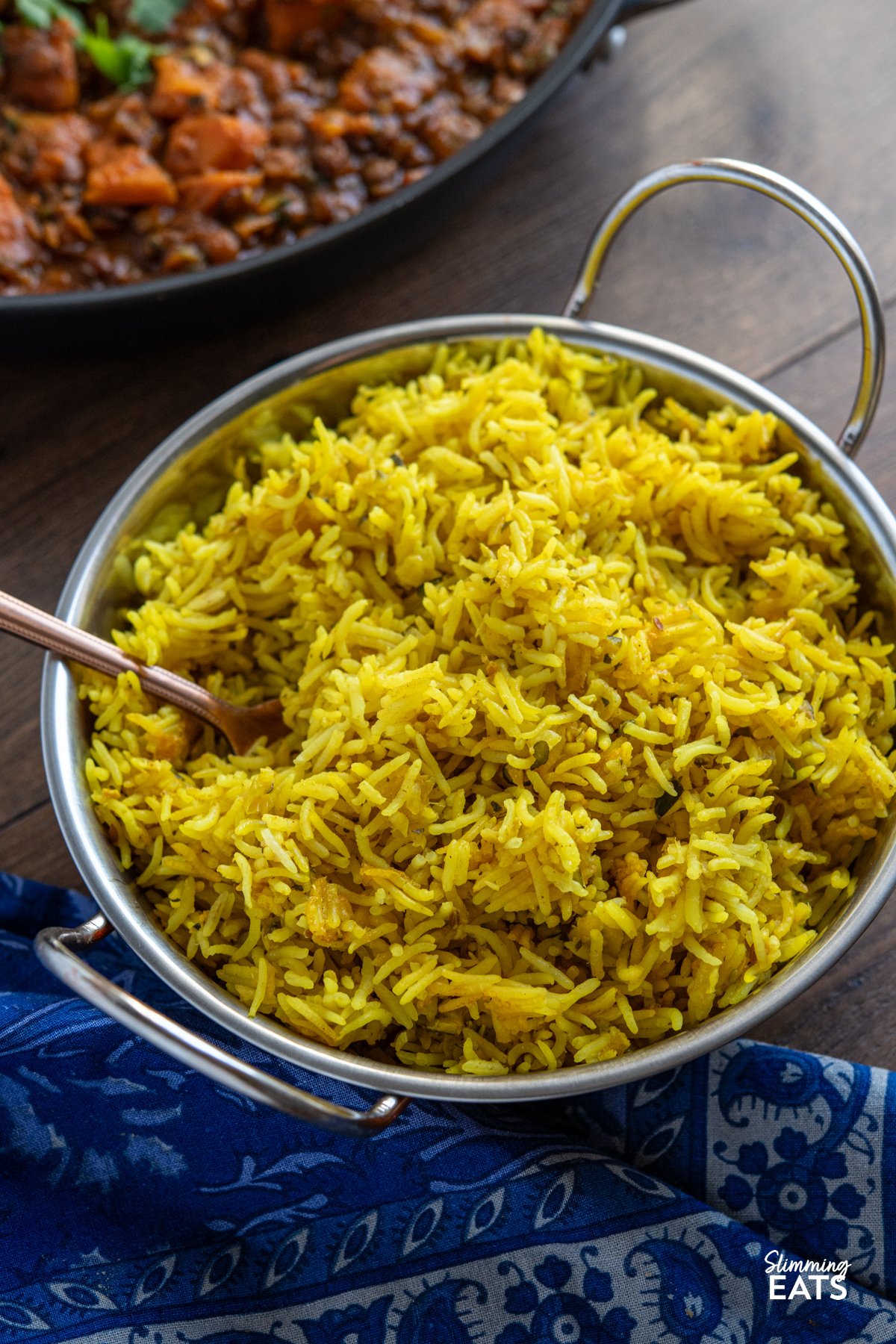 Zucchini Pilau Rice is a fragrant blend of basmati rice, tender zucchini, and warming spices like cumin and turmeric. Light yet flavourful, it’s perfect for a quick dinner or as a vibrant side dish. zucchini pilau rice in a double handled metal dish with serving spoon placed in rice, pan of curry visible in background