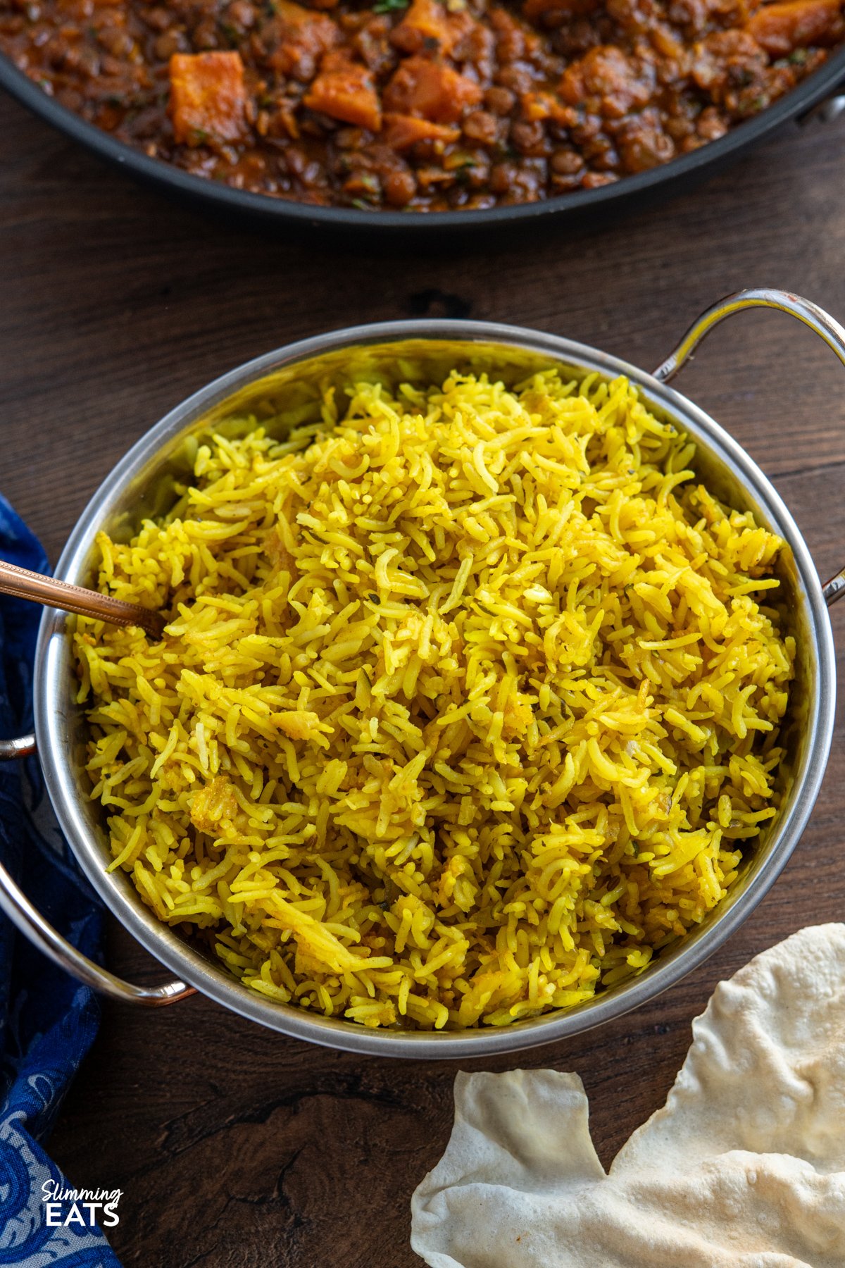 Zucchini Pilau Rice is a fragrant blend of basmati rice, tender zucchini, and warming spices like cumin and turmeric. Light yet flavourful, it’s perfect for a quick dinner or as a vibrant side dish. zucchini pilau rice in a double handled metal dish with serving spoon placed in rice, pan of curry visible in background