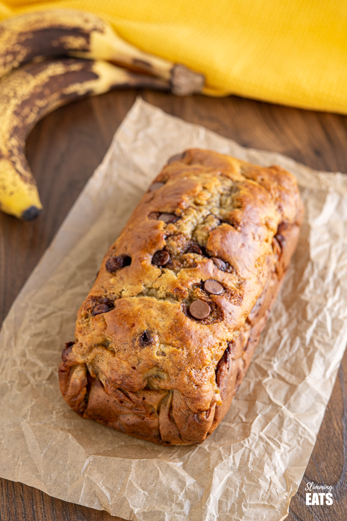 Healthy Banana and Chocolate Chip Loaf on parchment paper with ripe bananas in the background and yellow cloth