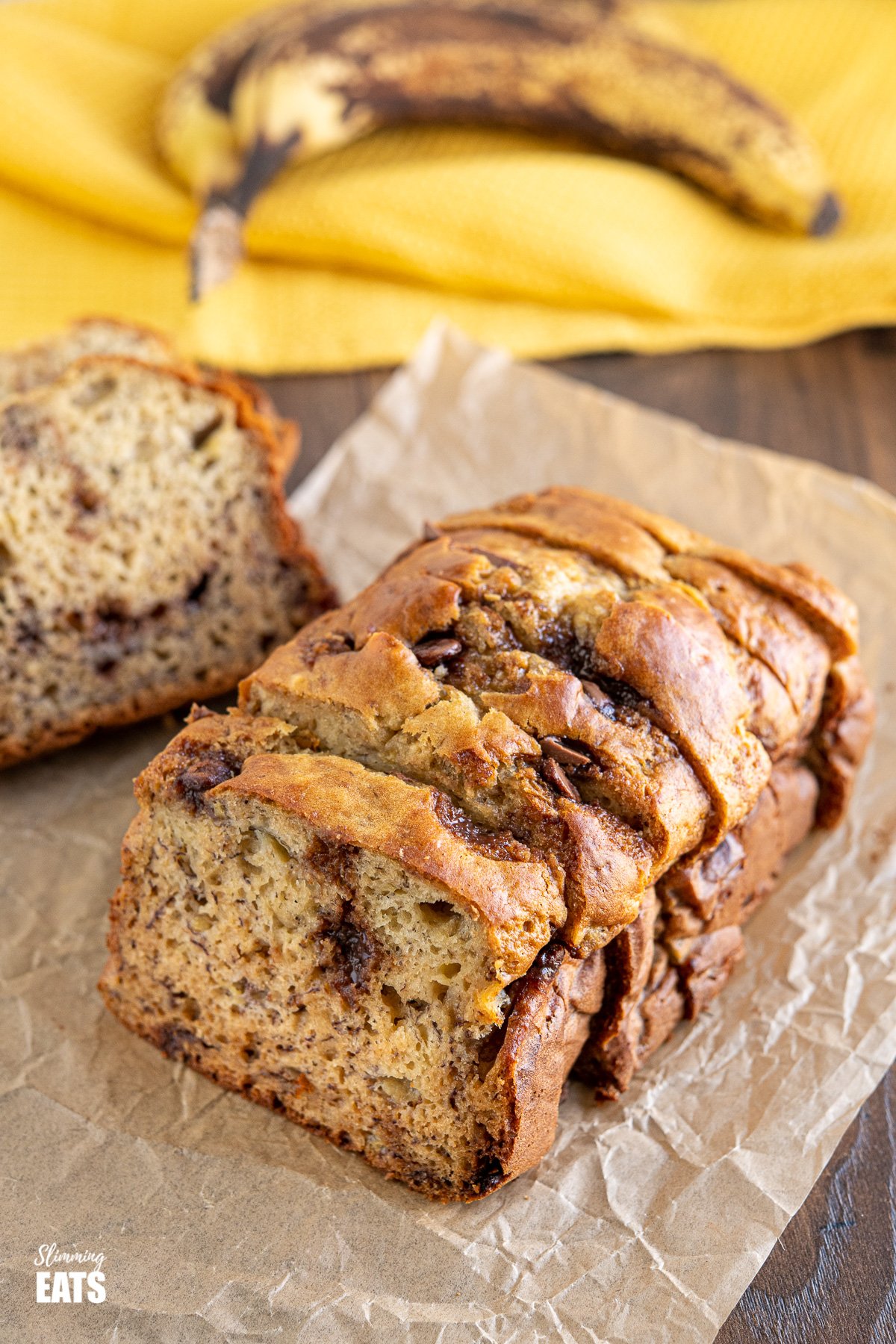 sliced Healthy Banana and Chocolate Chip Loaf on parchment paper with ripe bananas in background on yellow tea towel