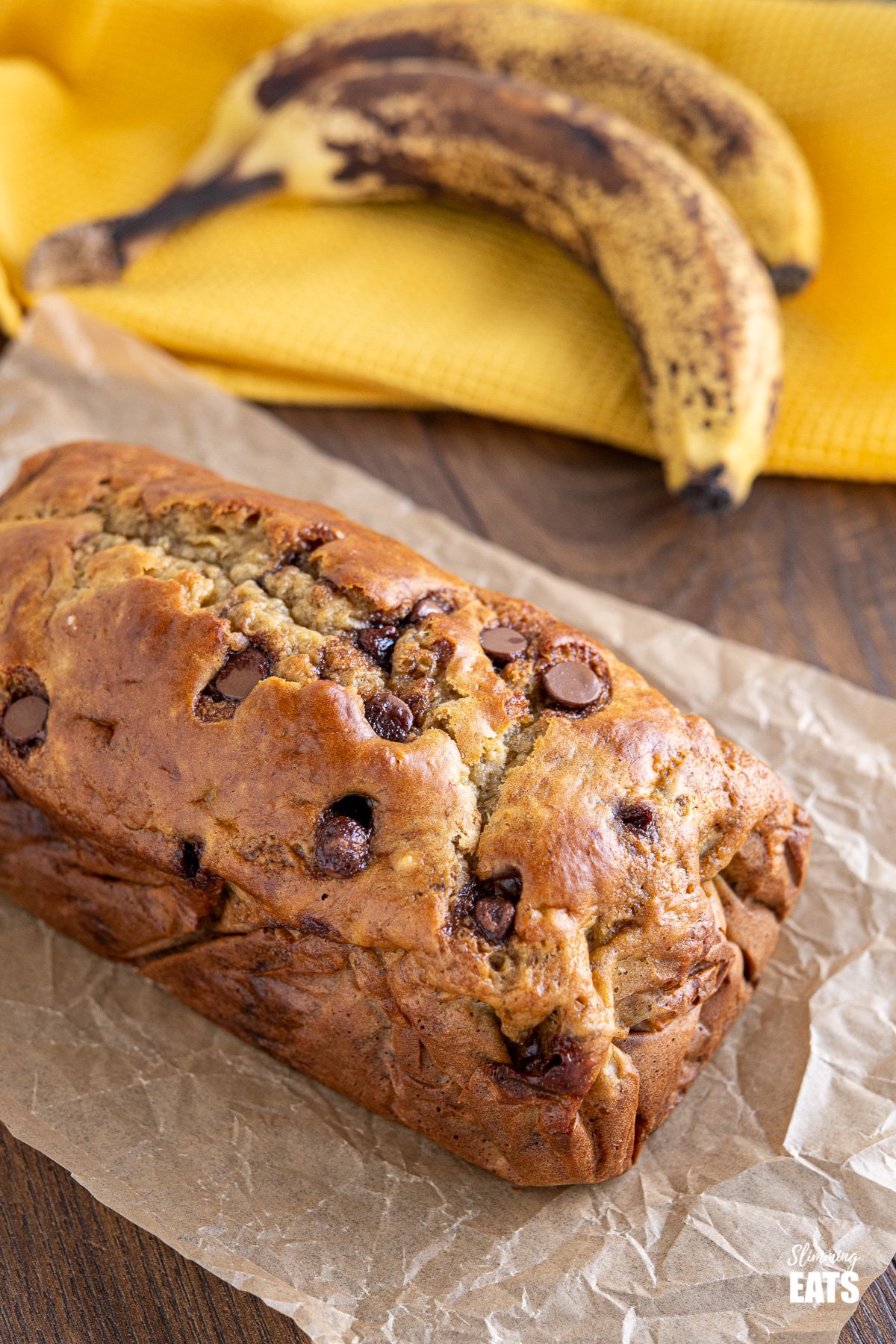 Healthy Banana and Chocolate Chip Loaf on parchment paper with ripe bananas in background on yellow tea towel