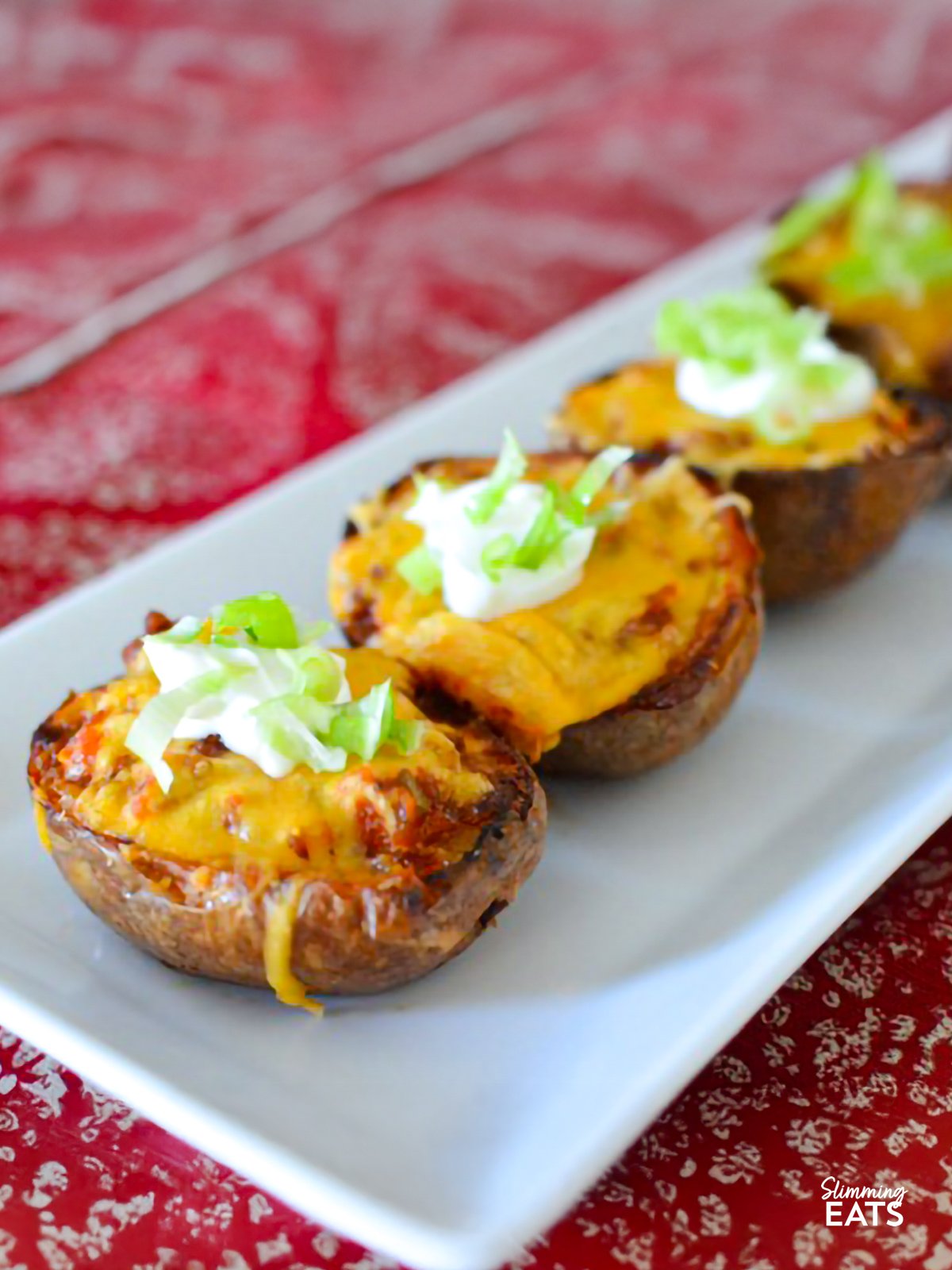 These Chilli Stuffed Potato Skins are a comforting and indulgent dish, with tender potato skins filled with flavour-packed chilli and topped with creamy soured cream. Perfect for a snack or a hearty meal, they’re sure to please everyone at the table. Chilli-Stuffed Potato Skins on a white rectangle plate place on a red patterned placemat