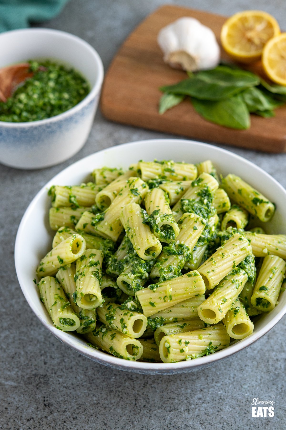 A delicious green pesto made from arugula, basil and spinach that is slightly lower on the calories, meaning you can pile this on a bowl of pasta by the spoonful and dig in a enjoy. #weightwatchers #lowcalorie skinny pesto on cooked pasted in a white bowl with basil, lemon and garlic, and bowl of homemade pesto behind