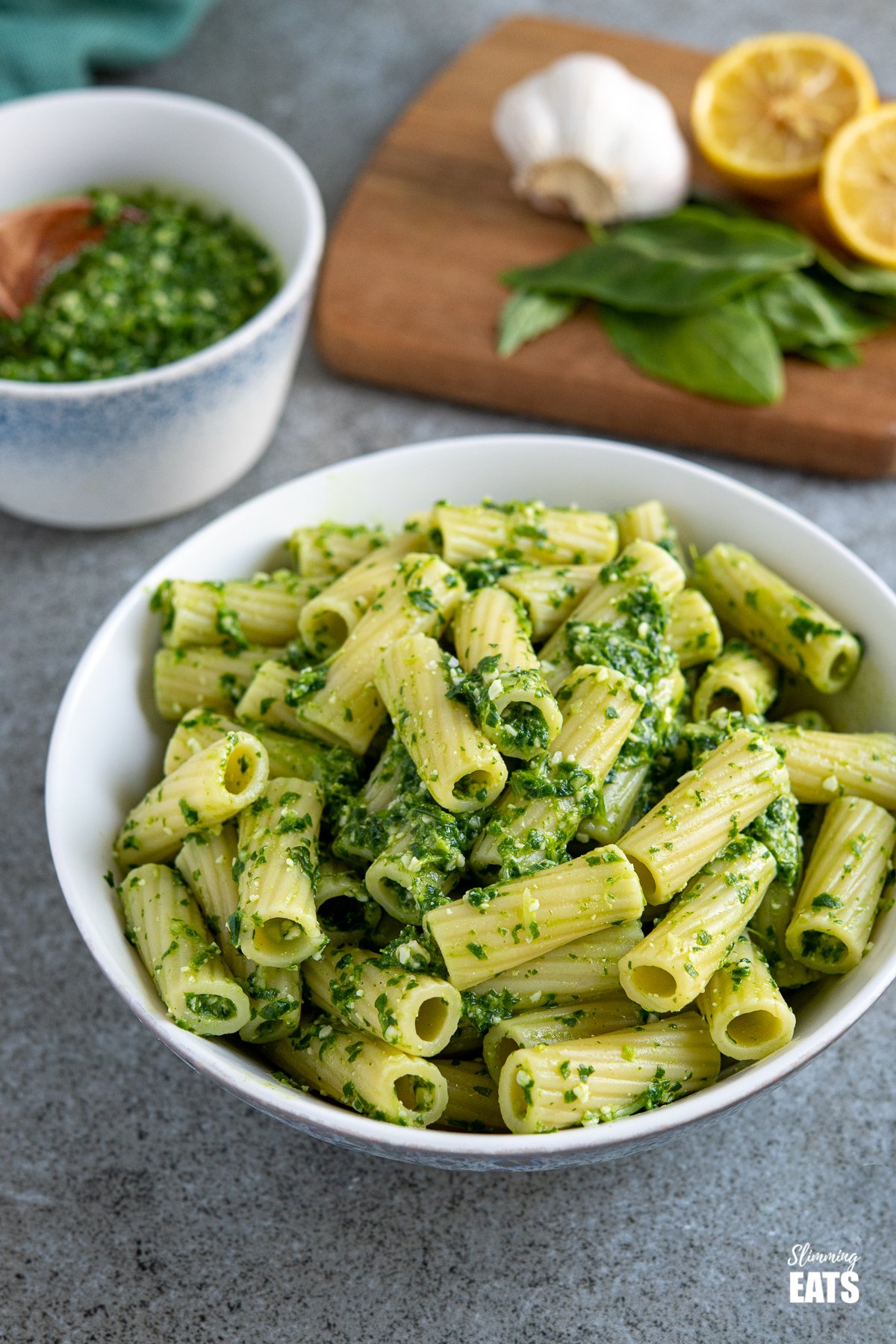 skinny pesto on cooked pasted in a white bowl with basil, lemon and garlic, and bowl of homemade pesto behind