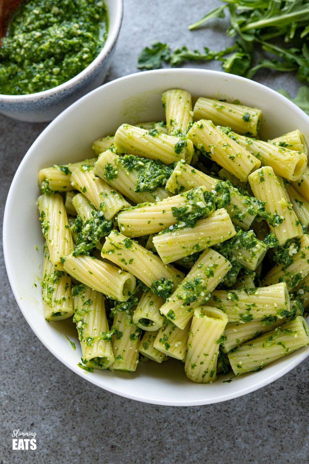 skinny pesto on cooked pasted in a white bowl with scattered arugula and bowl of homemade pesto behind