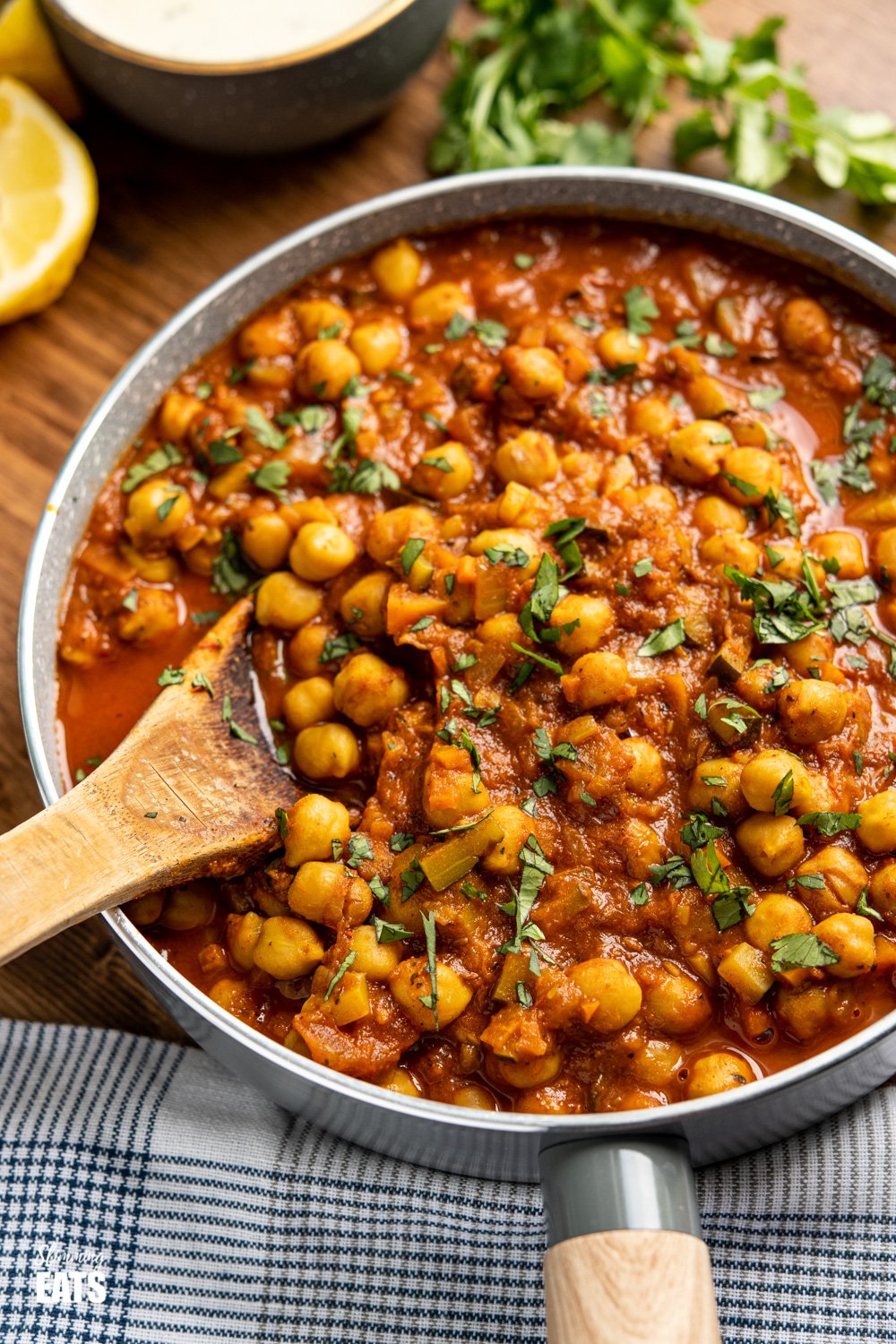 Chana Masala in a pan with wooden spoon, coriander and lemon wedges in the background on a wooden board. 