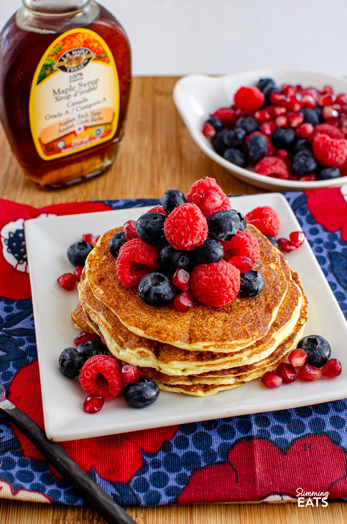 High Protein Cottage Cheese Pancakes served on a white plate, garnished with a variety of mixed berries and a light drizzle of maple syrup, with a bottle of maple syrup and additional berries in the background.