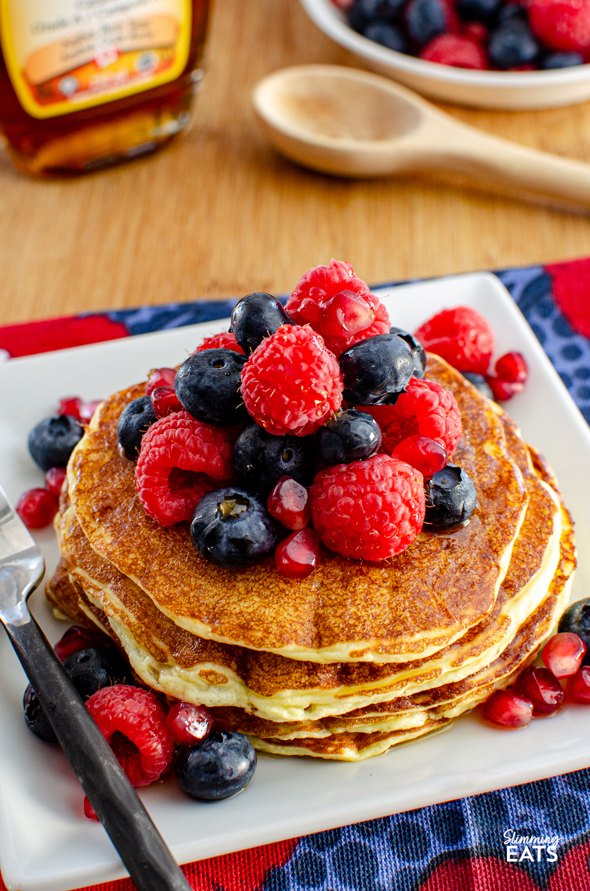 High Protein Cottage Cheese Pancakes served on a white plate, garnished with a variety of mixed berries and a light drizzle of maple syrup, with a bottle of maple syrup and additional berries in the background.