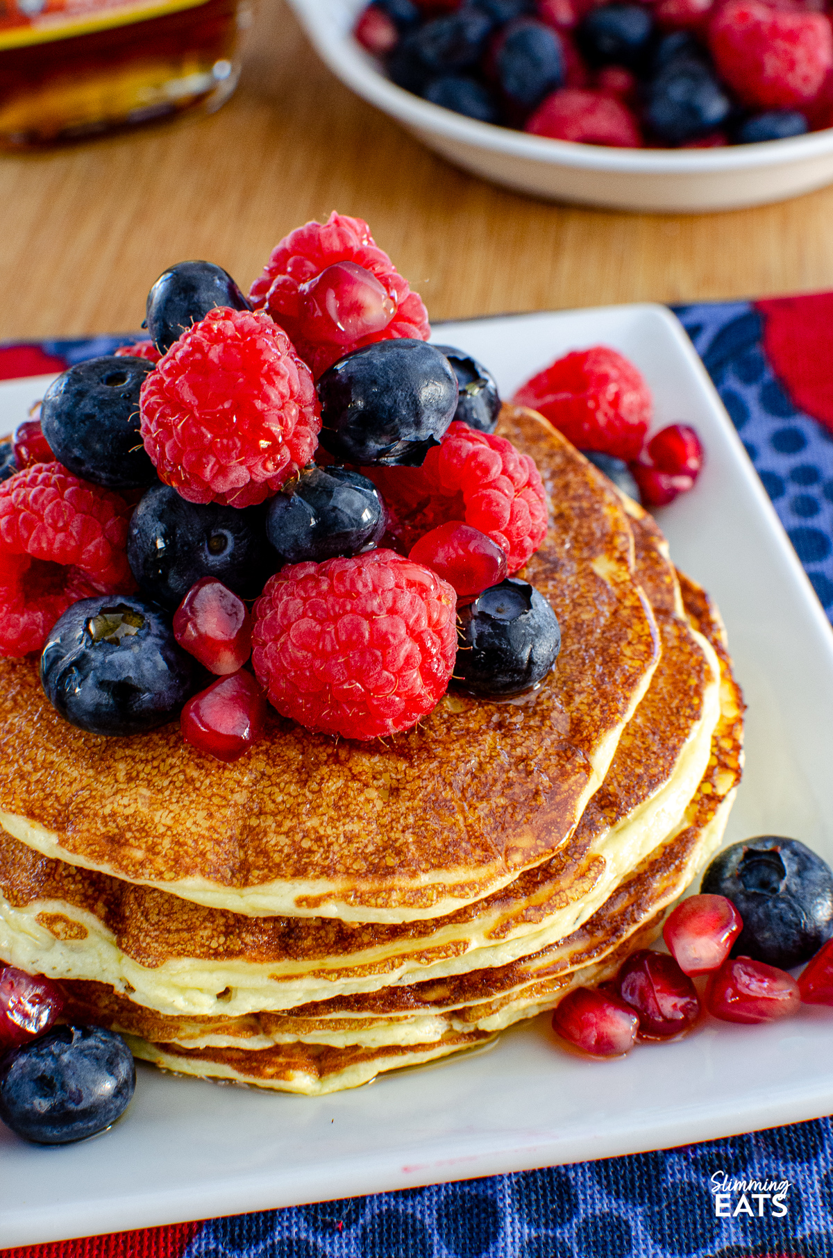 High Protein Cottage Cheese Pancakes served on a white plate, garnished with a variety of mixed berries and a light drizzle of maple syrup, with a bottle of maple syrup and additional berries in the background.