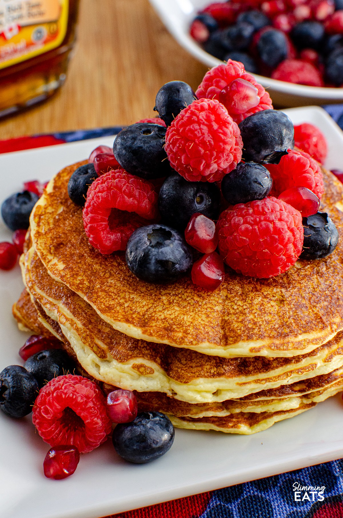 High Protein Cottage Cheese Pancakes served on a white plate, garnished with a variety of mixed berries and a light drizzle of maple syrup, with a bottle of maple syrup and additional berries in the background.