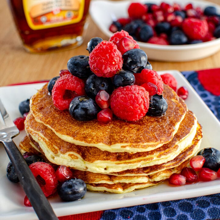 High Protein Cottage Cheese Pancakes served on a white plate, garnished with a variety of mixed berries and a light drizzle of maple syrup, with a bottle of maple syrup and additional berries in the background.