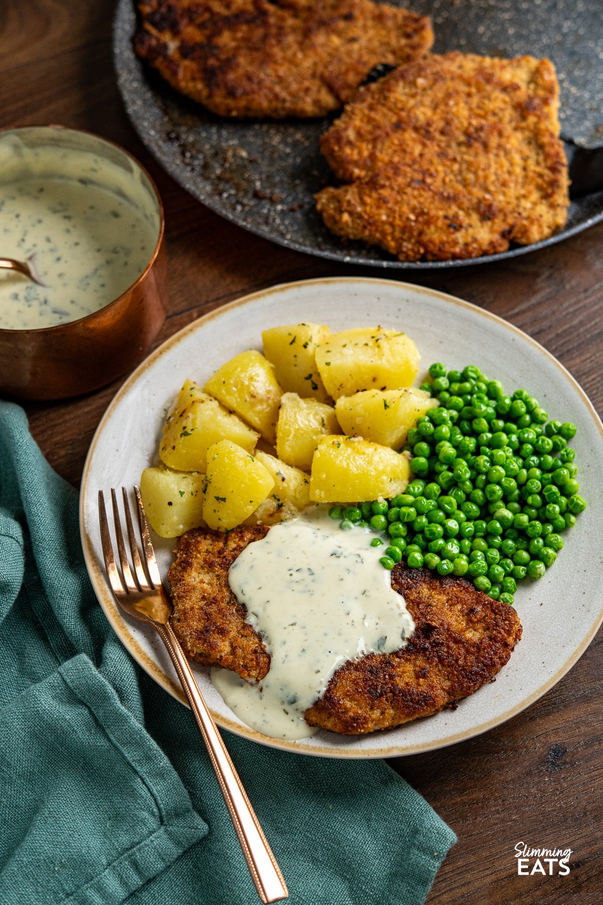 Indulge in a family-favourite pork schnitzel, featuring tender cutlets coated in seasoned wholemeal breadcrumbs, pan-fried to golden perfection, and served with a delicious creamy parsley sauce. Pork Schnitzel on plate drizzled with parsley sauce with lemon potatoes and garden peas