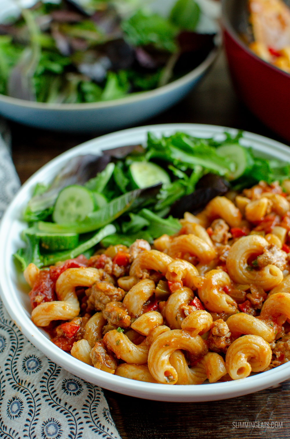 sausage and tomato pasta in white bowl with mixed baby greens and cucumber