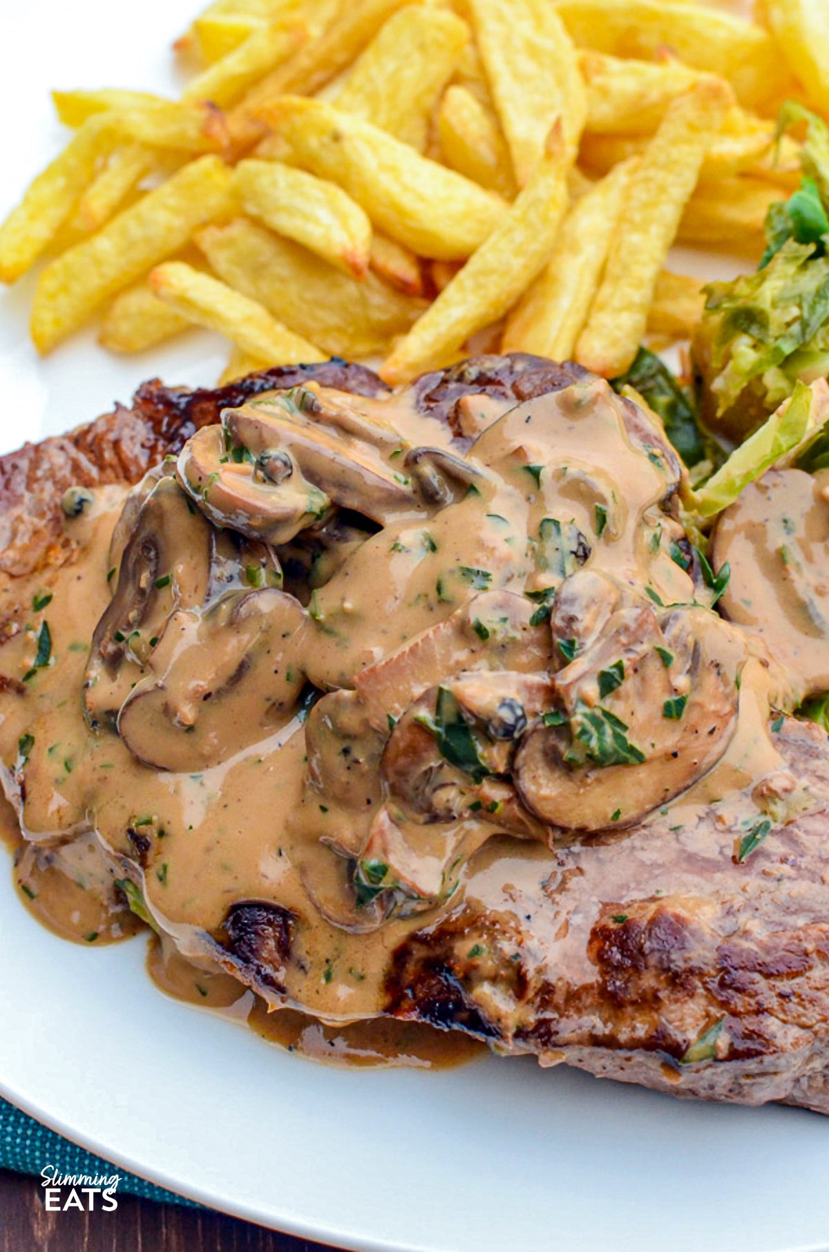 Steak with creamy peppercorn sauce served on a white plate alongside homemade oven-baked fries and sautéed Brussels sprouts.