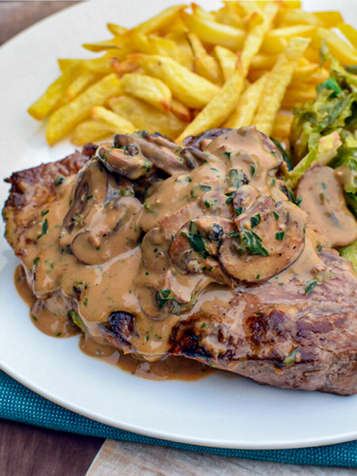 Steak with creamy peppercorn sauce served on a white plate alongside homemade oven-baked fries and sautéed Brussels sprouts