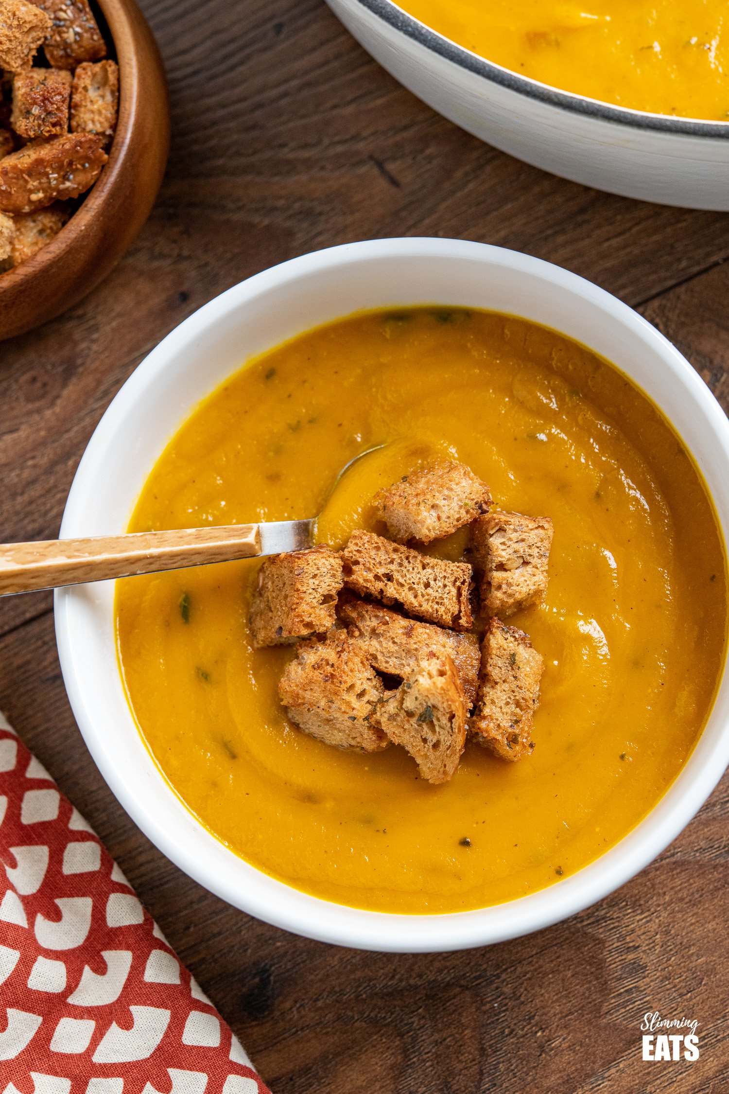 over the top view of Butternut Squash soup with Garlic Herb Croutons in white bowl, with wooden bowl of croutons and pan of soup in background