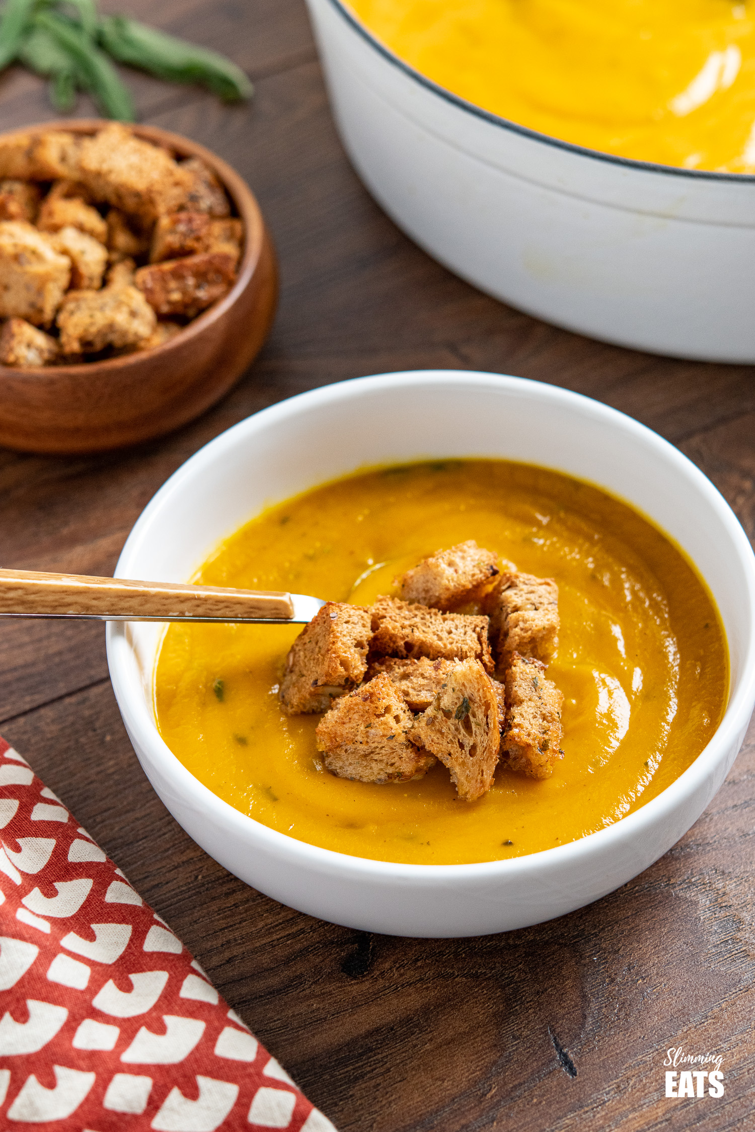 Butternut Squash soup with Garlic Herb Croutons in white bowl with wooden handles spoon and wooden bowl of croutons in background and pot of soup and scattered fresh sage leaves