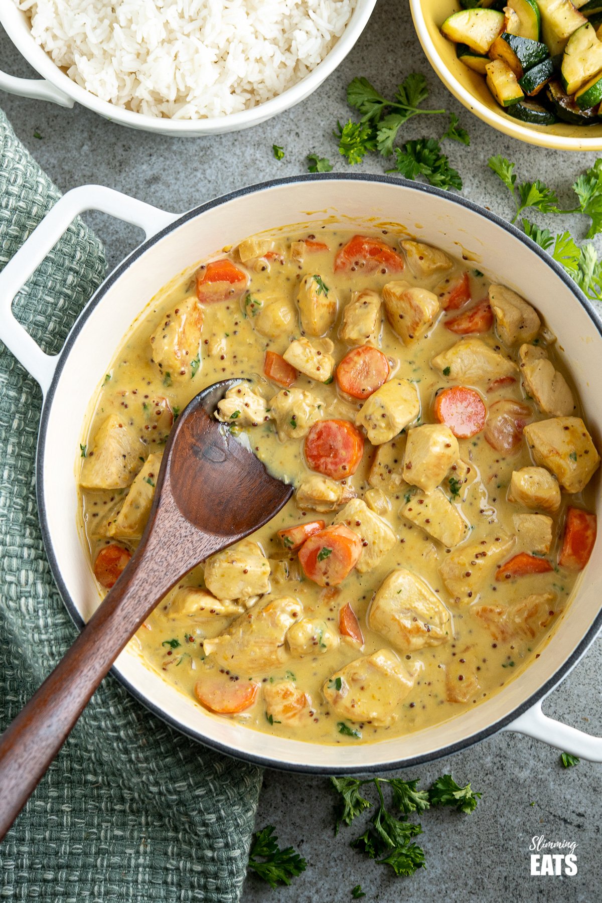Creamy Honey Mustard Chicken - tender chunks of chicken breast in a heavenly creamy honey and mustard sauce. #slimmingeats #weightwatchers #chicken #mustard #honey creamy honey mustard chicken in cast iron pan with dark wooden spoon, bowl of rice and veg in background and scattered parsley