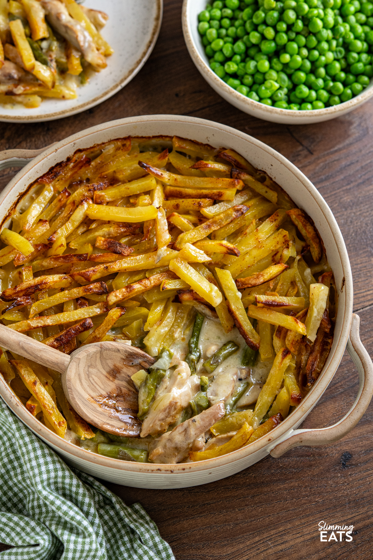 This Chicken and Asparagus Pie with Crispy Potatoes has a creamy, comforting filling topped with golden crispy potatoes for a delicious twist on a classic pie. Crispy Potato Topped Chicken and Asparagus Pie in ceramic oven dish with two handles surrounded by a bowl of garden peas and plate with serving of pie