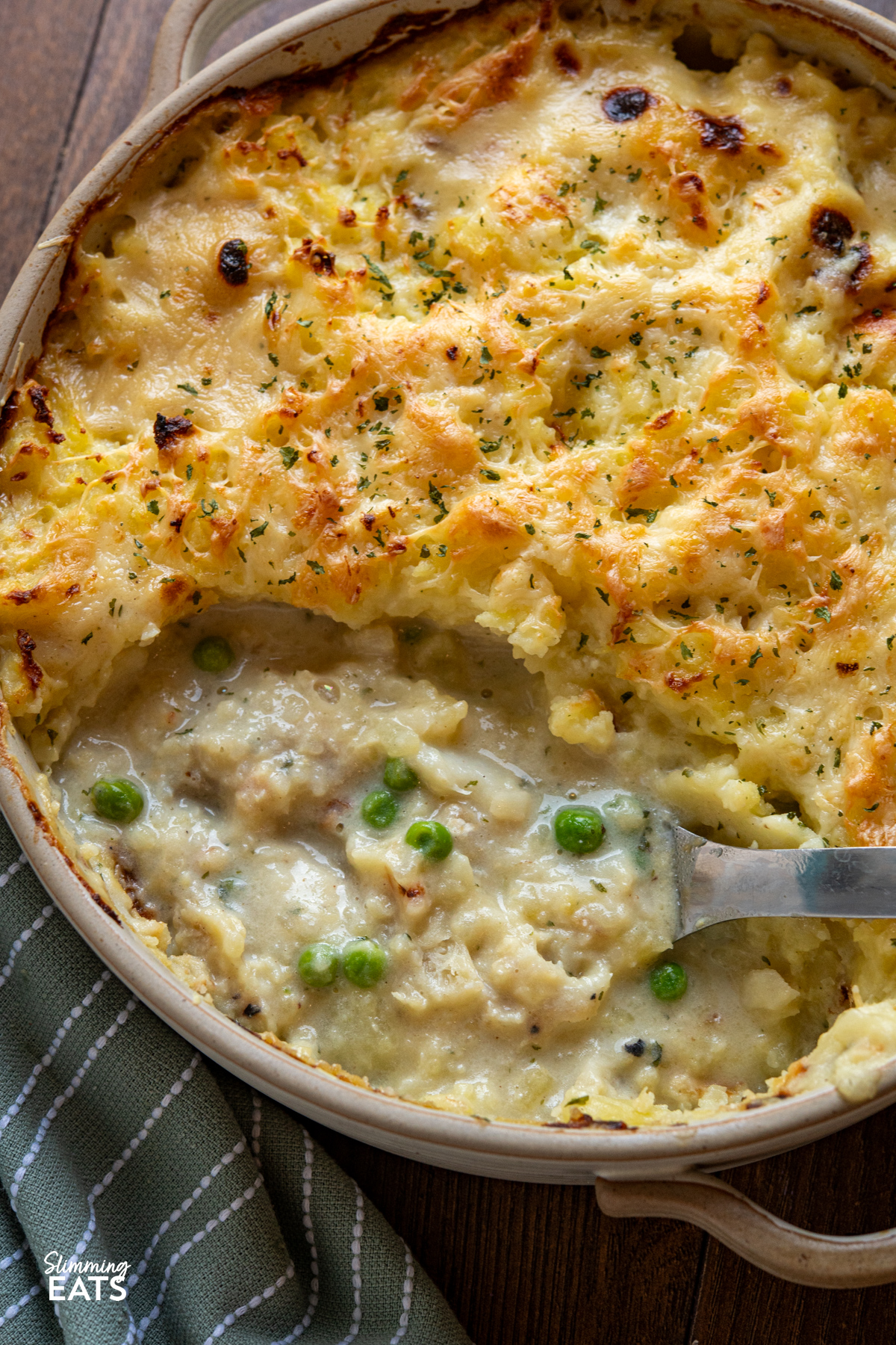 close up of Cheesy Topped Fish Pie in double handled beige round oven dish with serving spoon