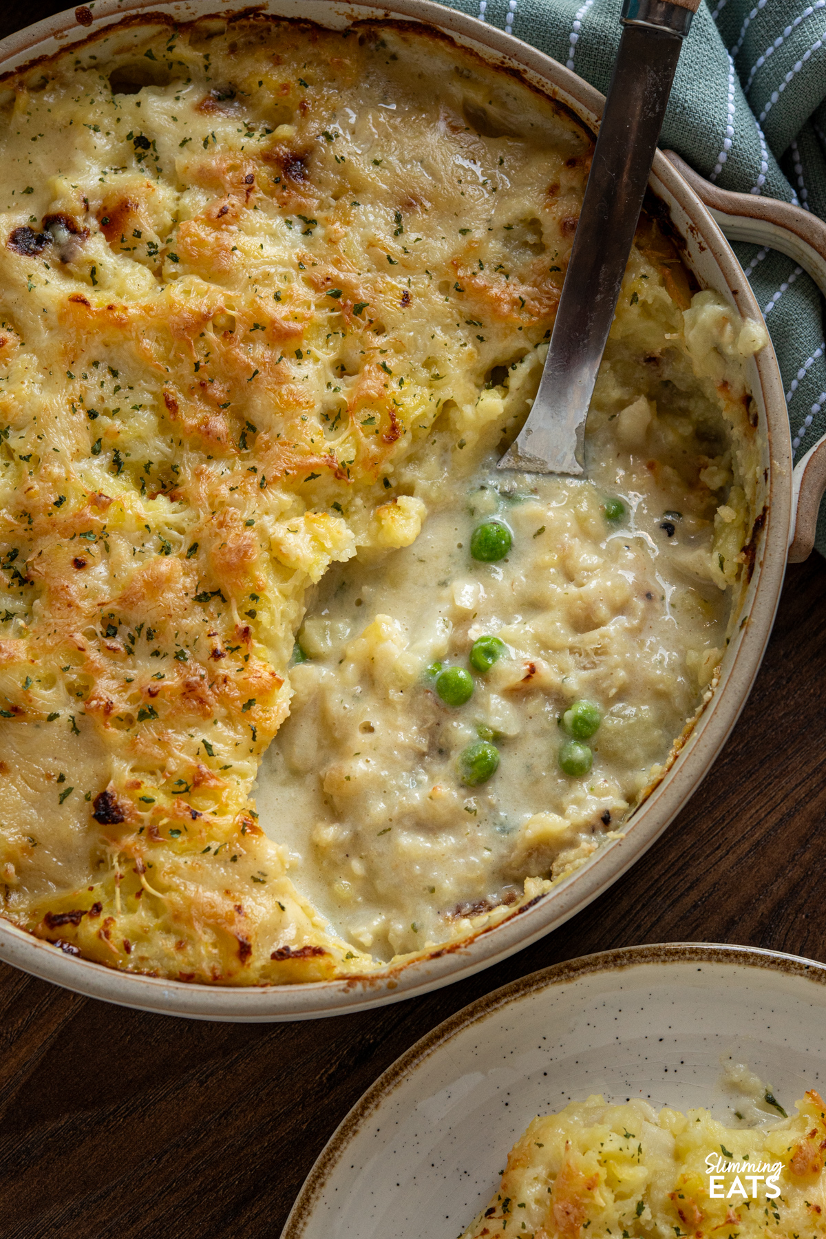 close up of Cheesy Topped Fish Pie in double handled beige round oven dish with serving spoon