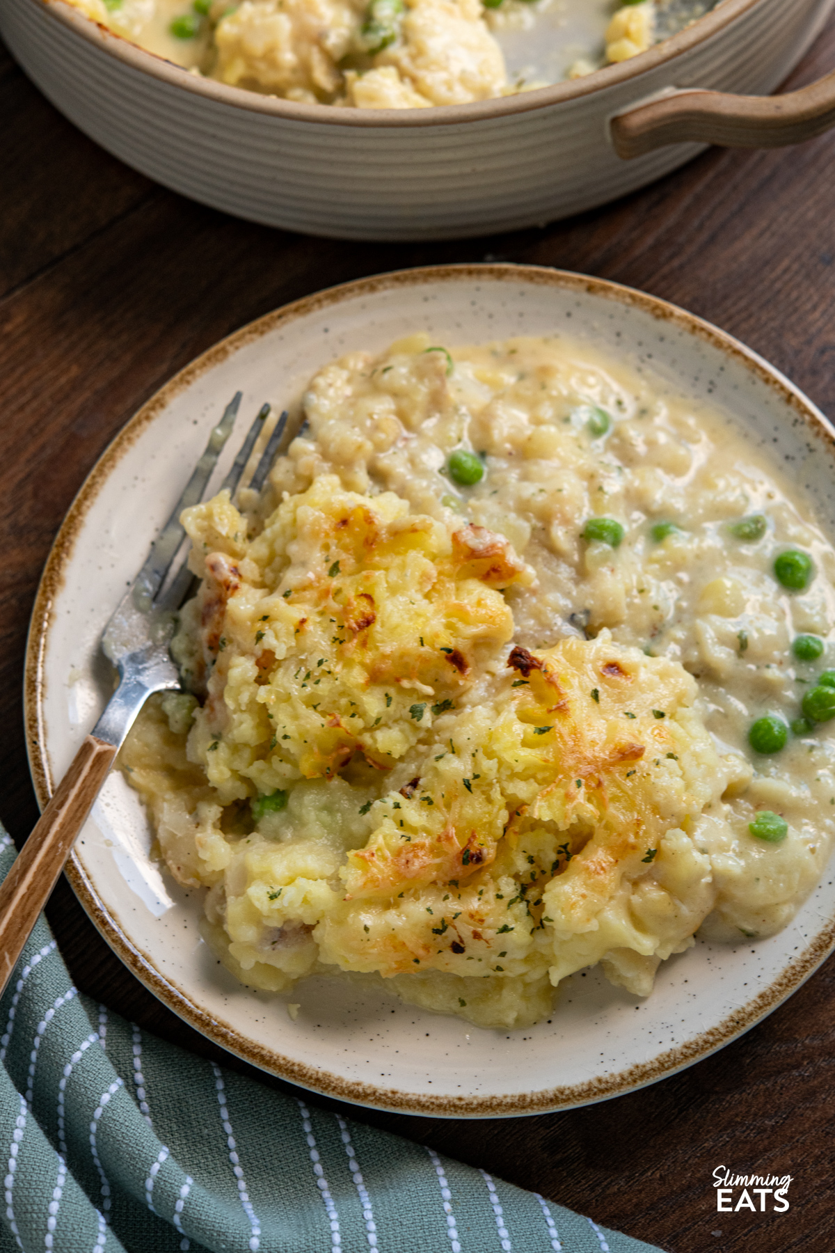 serving of cheesy fish pie on white plate with beige rim, fork place on left hand side
