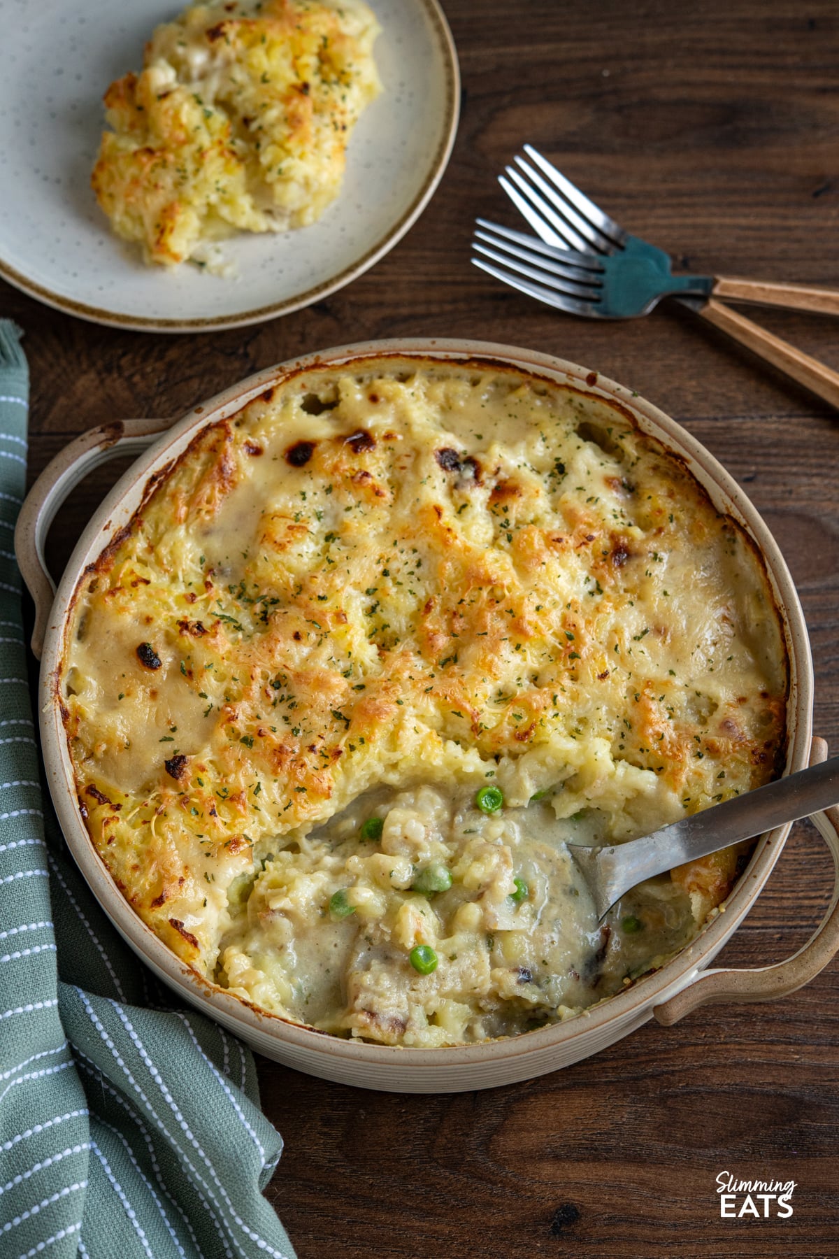 Cheesy Topped Fish Pie in double handled beige round oven dish, serving on plate with two forks above