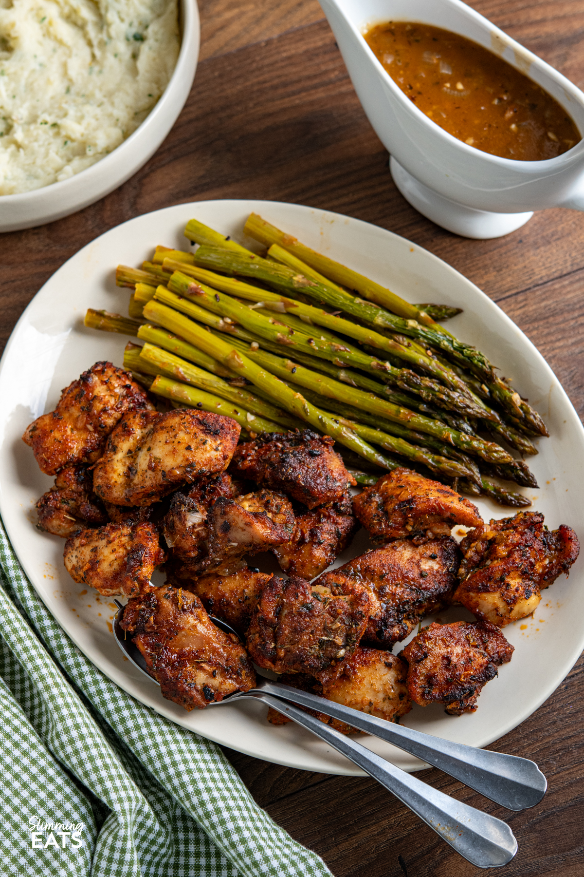 seasoned chicken, asparagus on a oval plate with spoon and fork, then jug with the sauce next to plate with spring onion mash