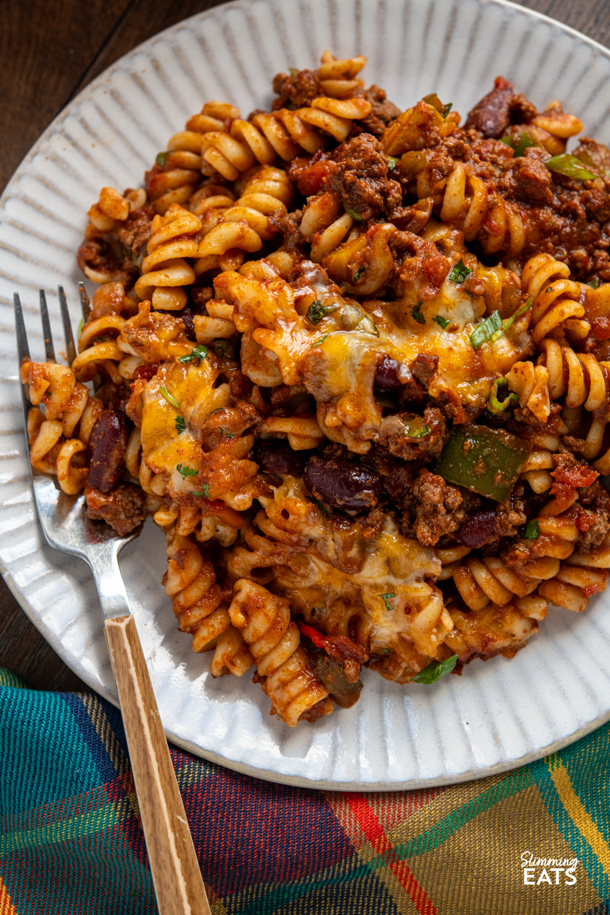 Mexican pasta bake is a hearty, comforting family meal that combines a rich beef chilli with pasta and a golden cheesy topping. close up of serving of Mexican pasta bake on a white ridged plate with fork place on left of plate