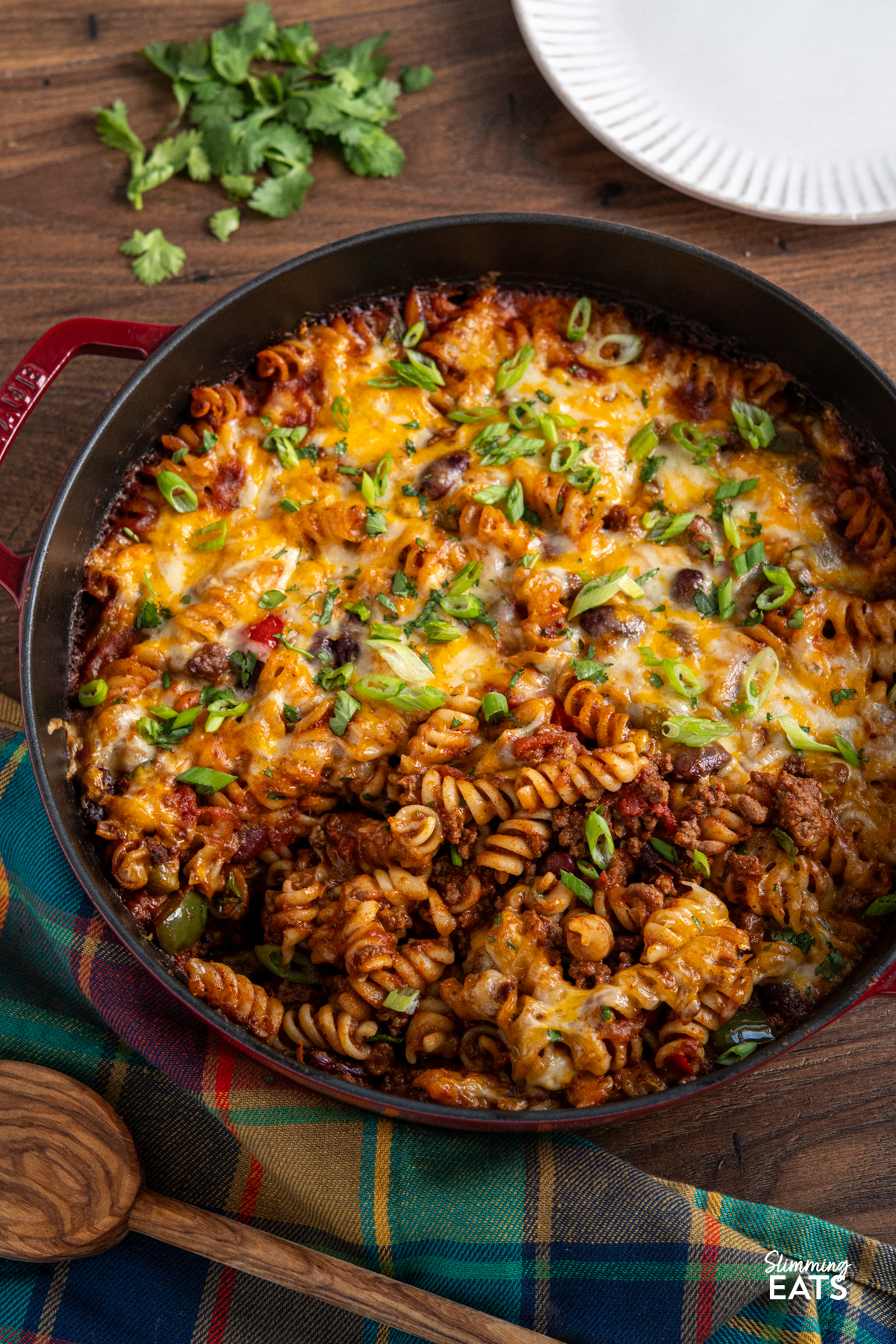 Mexican pasta bake is a hearty, comforting family meal that combines a rich beef chilli with pasta and a golden cheesy topping. Mexican pasta bake in a staub cherry cast iron double handled pan surrounded by cilantro, plate and wooden spoon