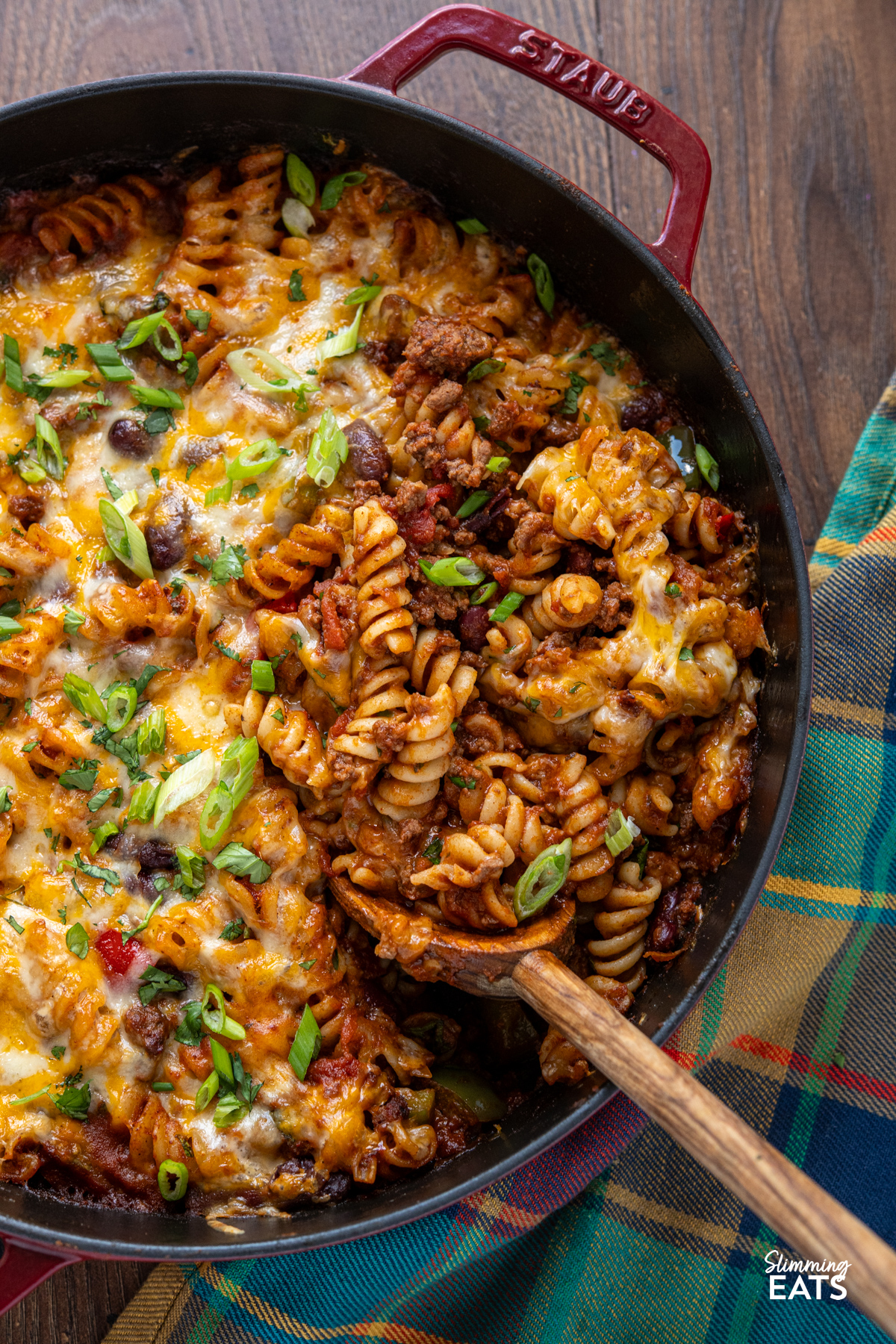 Mexican pasta bake is a hearty, comforting family meal that combines a rich beef chilli with pasta and a golden cheesy topping. close up of Mexican pasta bake in a staub cherry cast iron double handled pan
