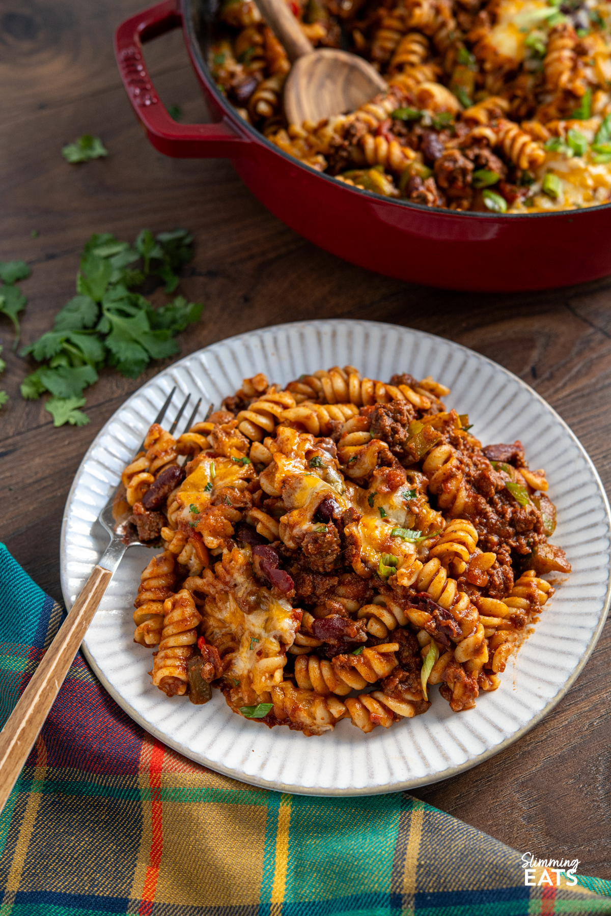 Mexican pasta bake is a hearty, comforting family meal that combines a rich beef chilli with pasta and a golden cheesy topping. serving of Mexican pasta bake on a white ridged plate with fork place on left of plate, cast iron pan with rest of mexican pasta bake visible