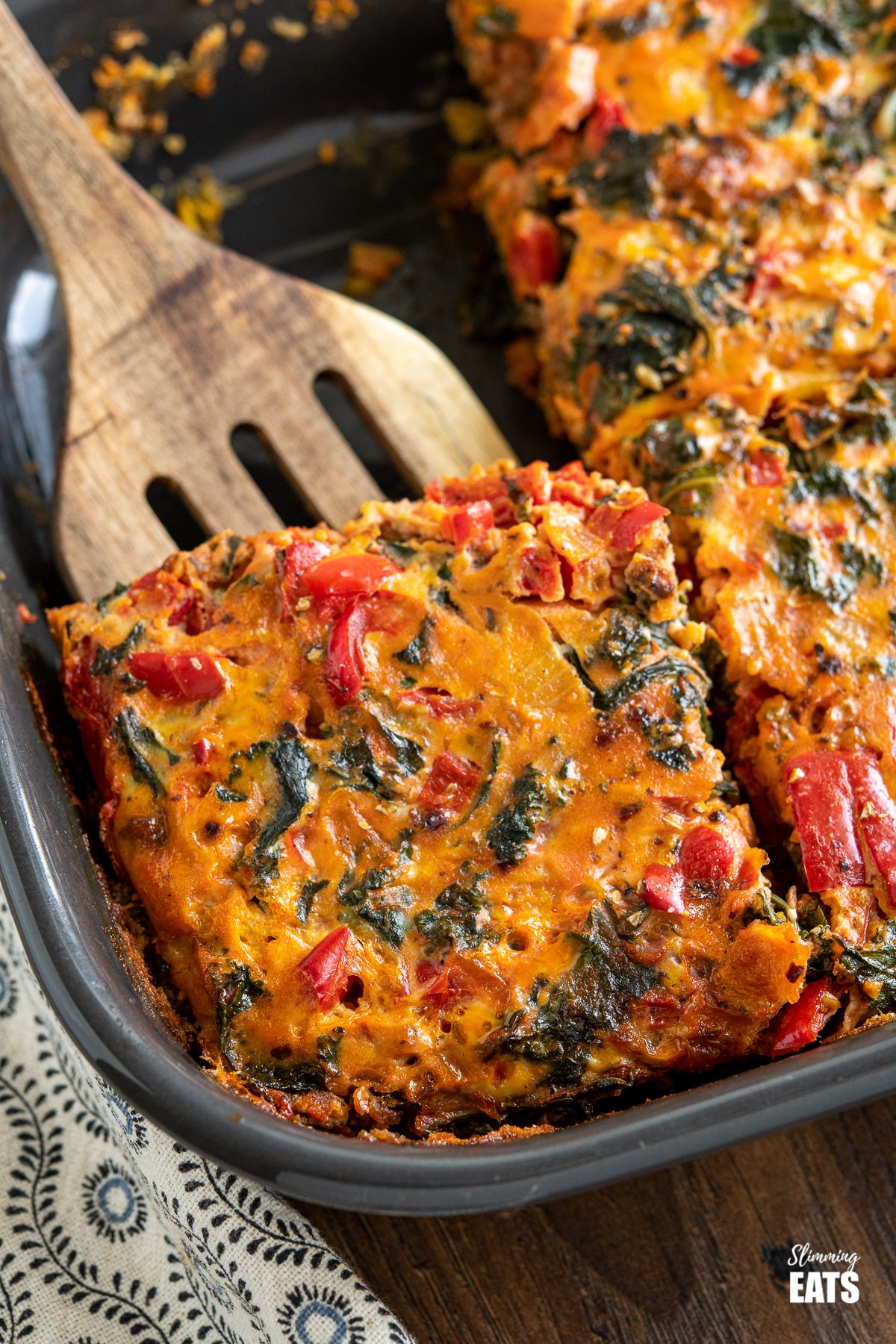 close up of a slice of Breakfast Layered Crustless Quiche in dark grey baking dish on wooden spatula