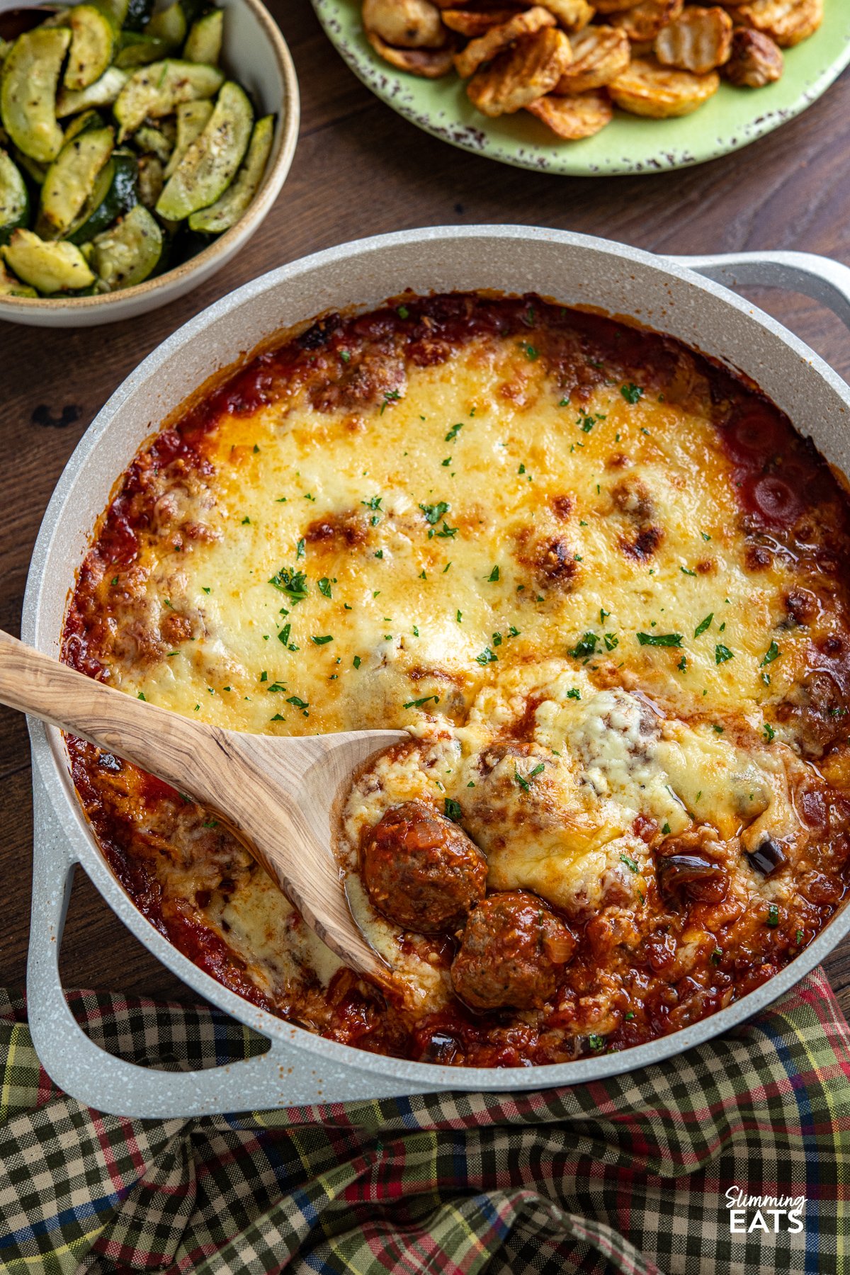 Indulge in the tantalizing flavors of the Mediterranean with this delicious creation of Moussaka Meatballs - tender seasoned lamb meatballs in a rich tomato sauce topped with a light creamy Bechamel sauce. Get ready to embark on a culinary journey that will transport your taste buds straight to the sun-soaked shores of Greece! Greek Moussaka Meatballs in a non stick ceramic casserole dish with potato and courgette sides